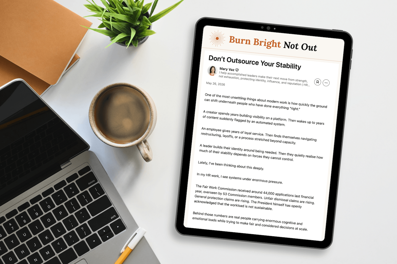 A desk with a silver laptop, a black tablet displaying an article titled 'Burn Bright Not Out' by Mary Vaz, a white coffee mug with coffee, a small green plant, and a closed brown notebook with a yellow pencil resting on it.