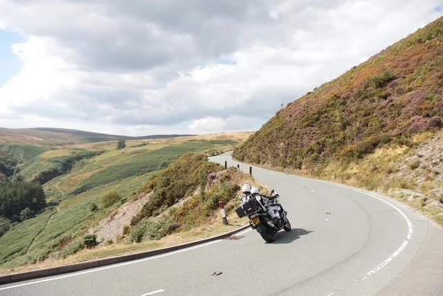 Motorcycle leaning into a curve on a mountain road with lush hills and cloudy sky in the background.