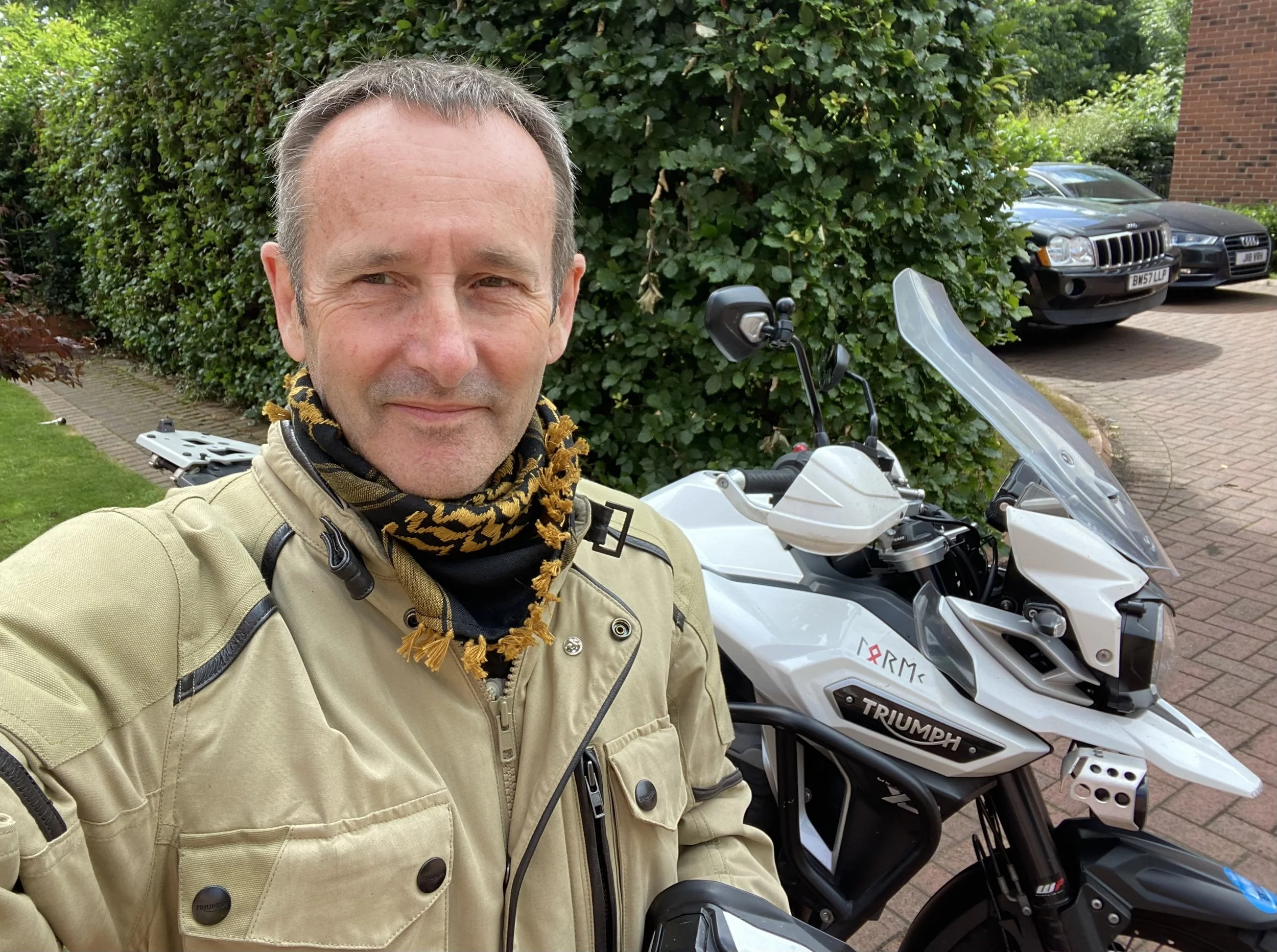 Man taking a selfie outdoors, standing next to a white motorcycle with a windshield. In the background are green bushes, two parked cars, and part of a brick building.