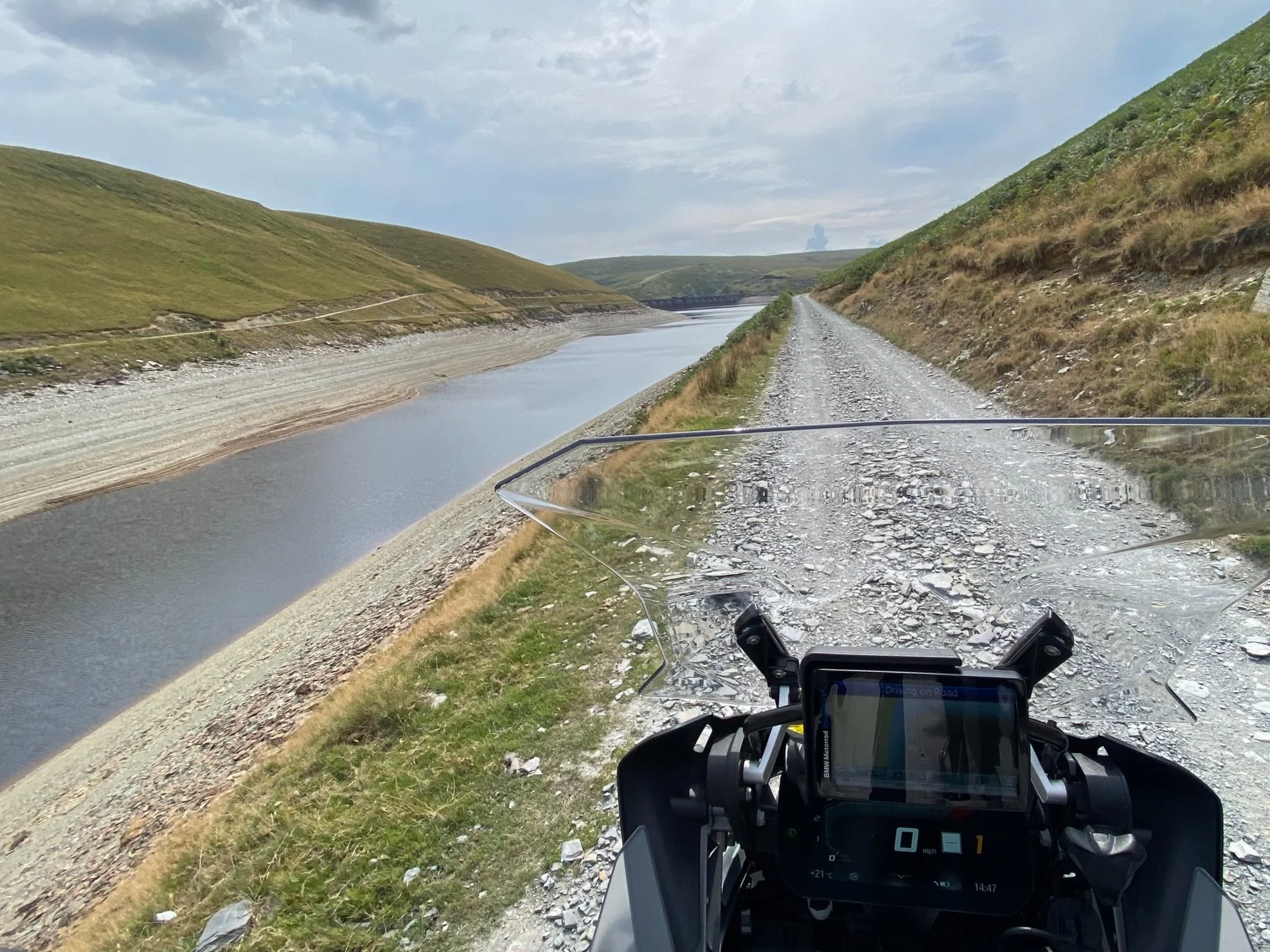 A gravel road runs alongside a calm river with green hills on either side under a partly cloudy sky, as seen from a motorcycle dashboard's perspective.