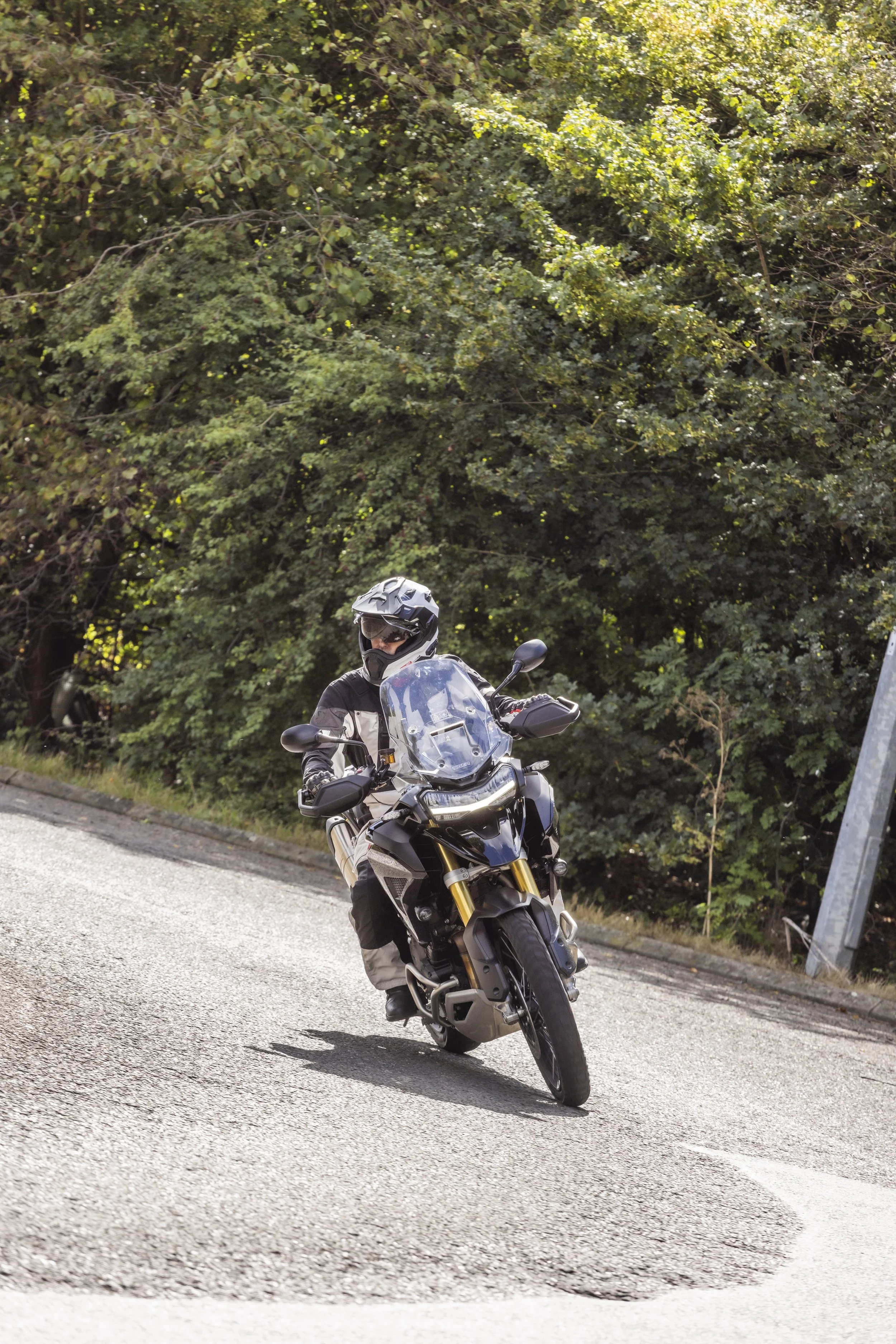 A motorcyclist wearing a helmet and riding gear, riding a black adventure motorcycle on a paved road with dense green trees in the background.