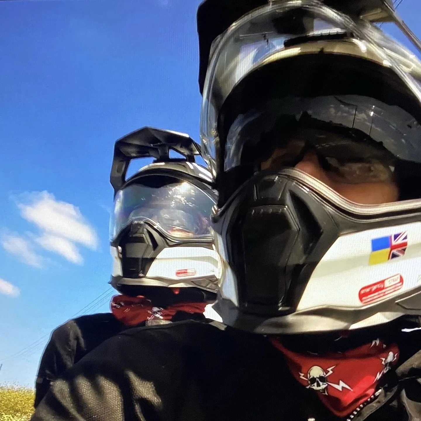 A group of motorcyclists with helmets, standing outdoors on a clear day with a blue sky and a few clouds, possibly on a ride or gathering.