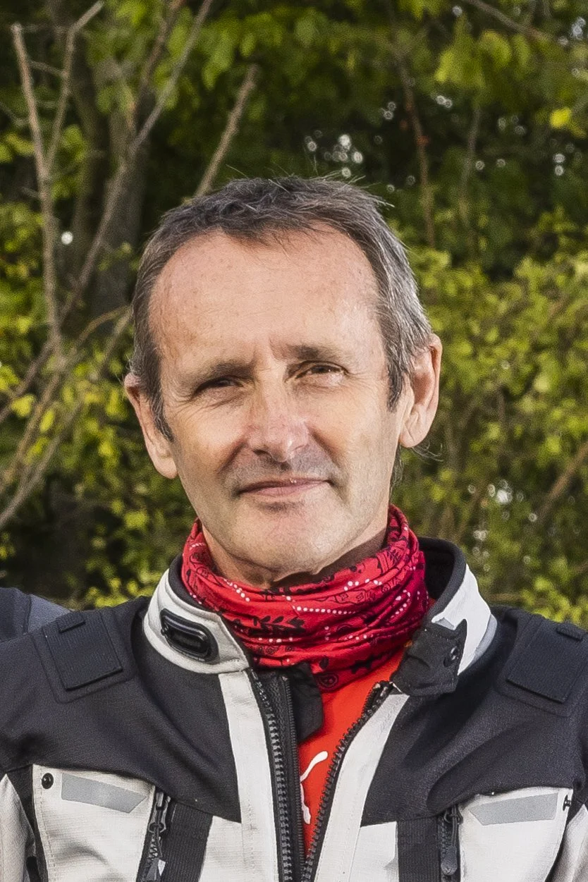 Man with brown hair wearing a black and white jacket, red neck gaiter, standing outdoors with trees and greenery in the background.