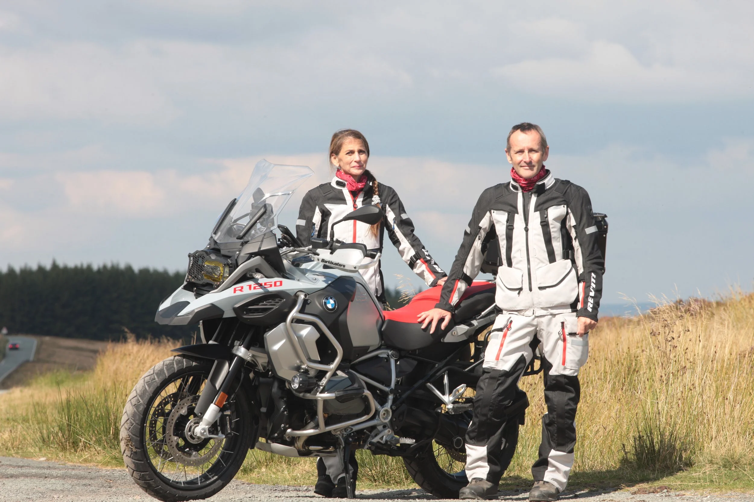 A man and a woman dressed in motorcycle riding gear standing outdoors beside a BMW R1250 GS motorcycle, with fields and a cloudy sky in the background.