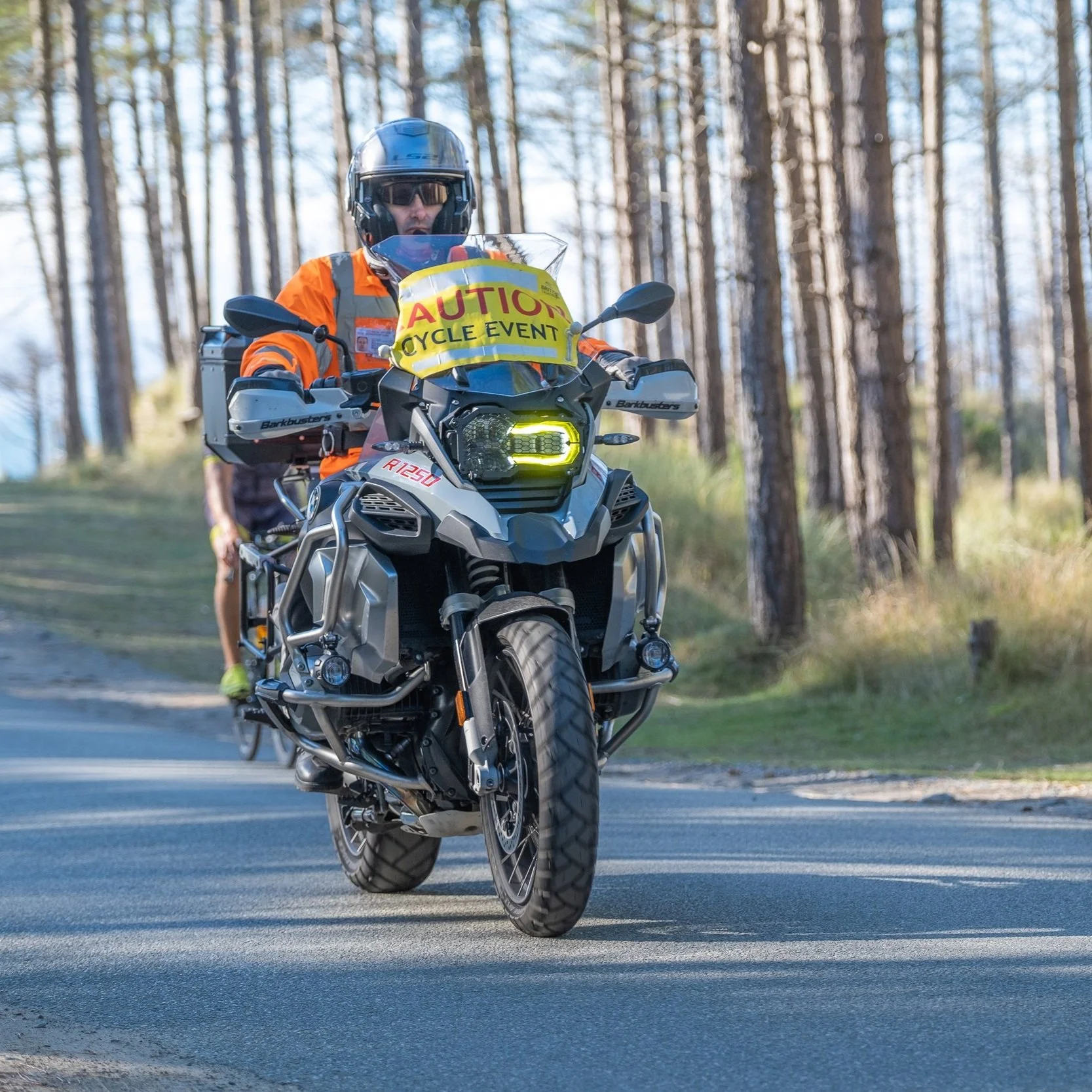 A person riding a gray motorcycle on a paved road in a wooded area, wearing a black helmet and orange jacket. The motorcycle has a yellow sign that reads "CAUTION" and "CYCLE EVENT."