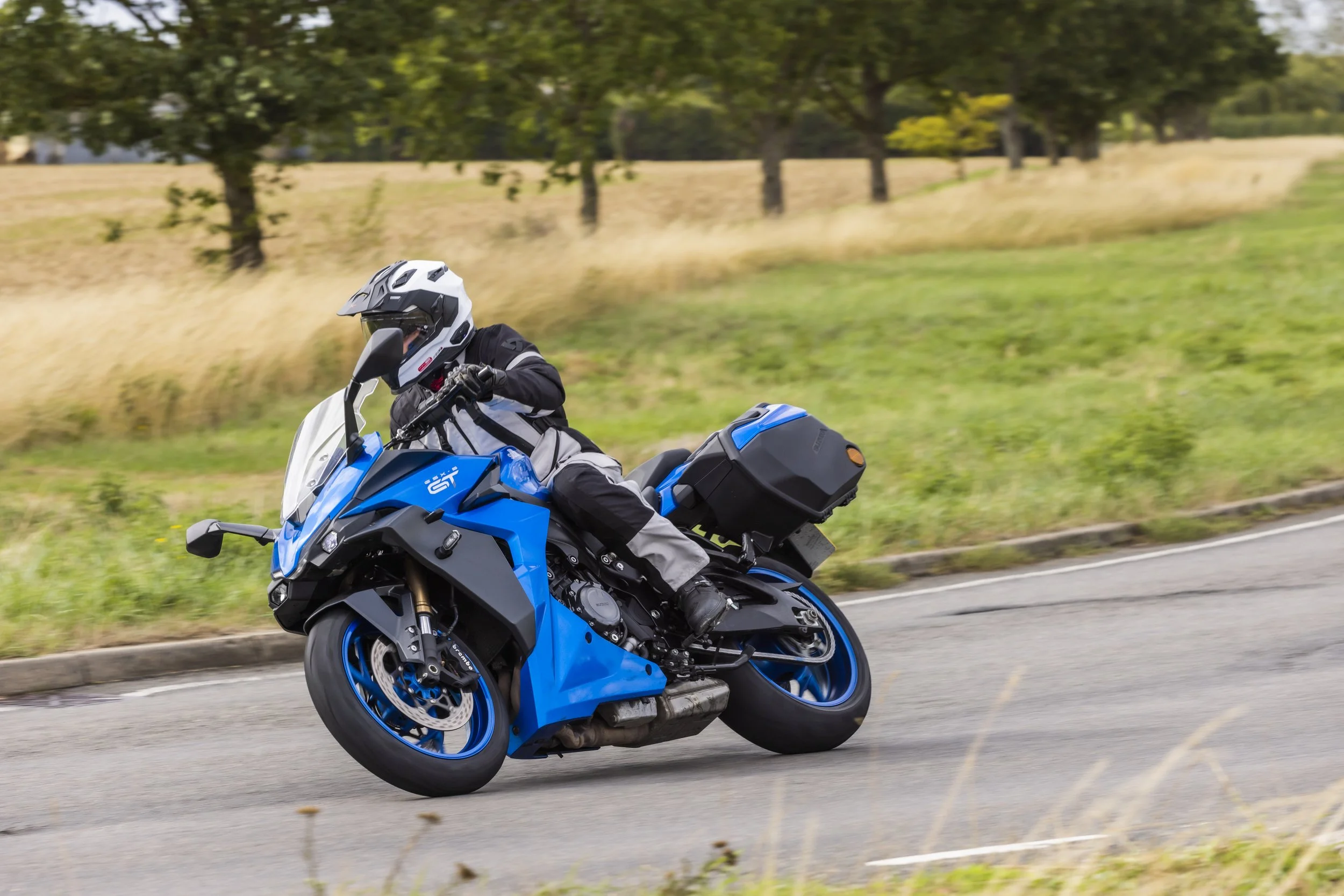 Motorcycle rider in black ridinggear and white helmet riding a blue motorcycle on a paved road with a grassy field and trees in the background.