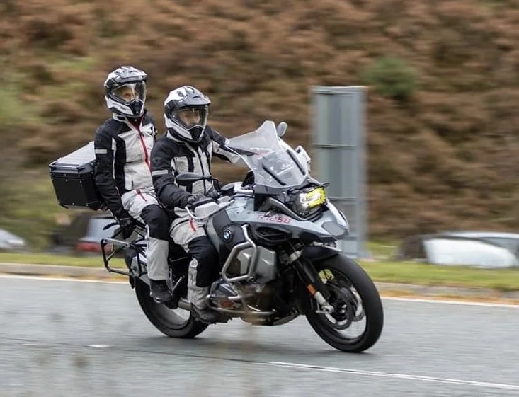 Two motorcyclists wearing helmets and protective gear riding a gray adventure motorcycle with a top box on a road.