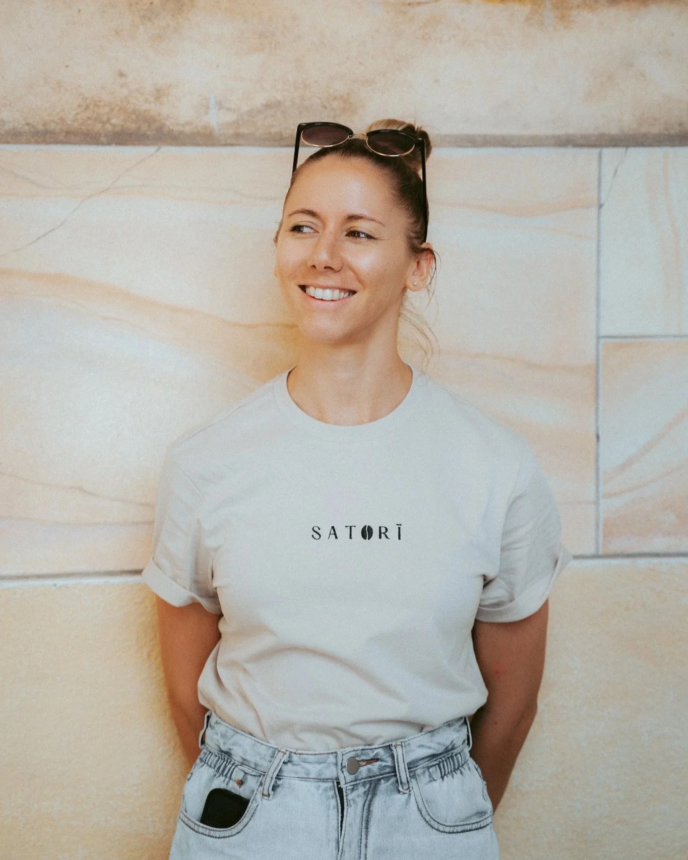 A woman leaning against a beige marble wall, wearing a light-colored T-shirt with the word 'SATORI' on it, sunglasses on her head, and smiling.