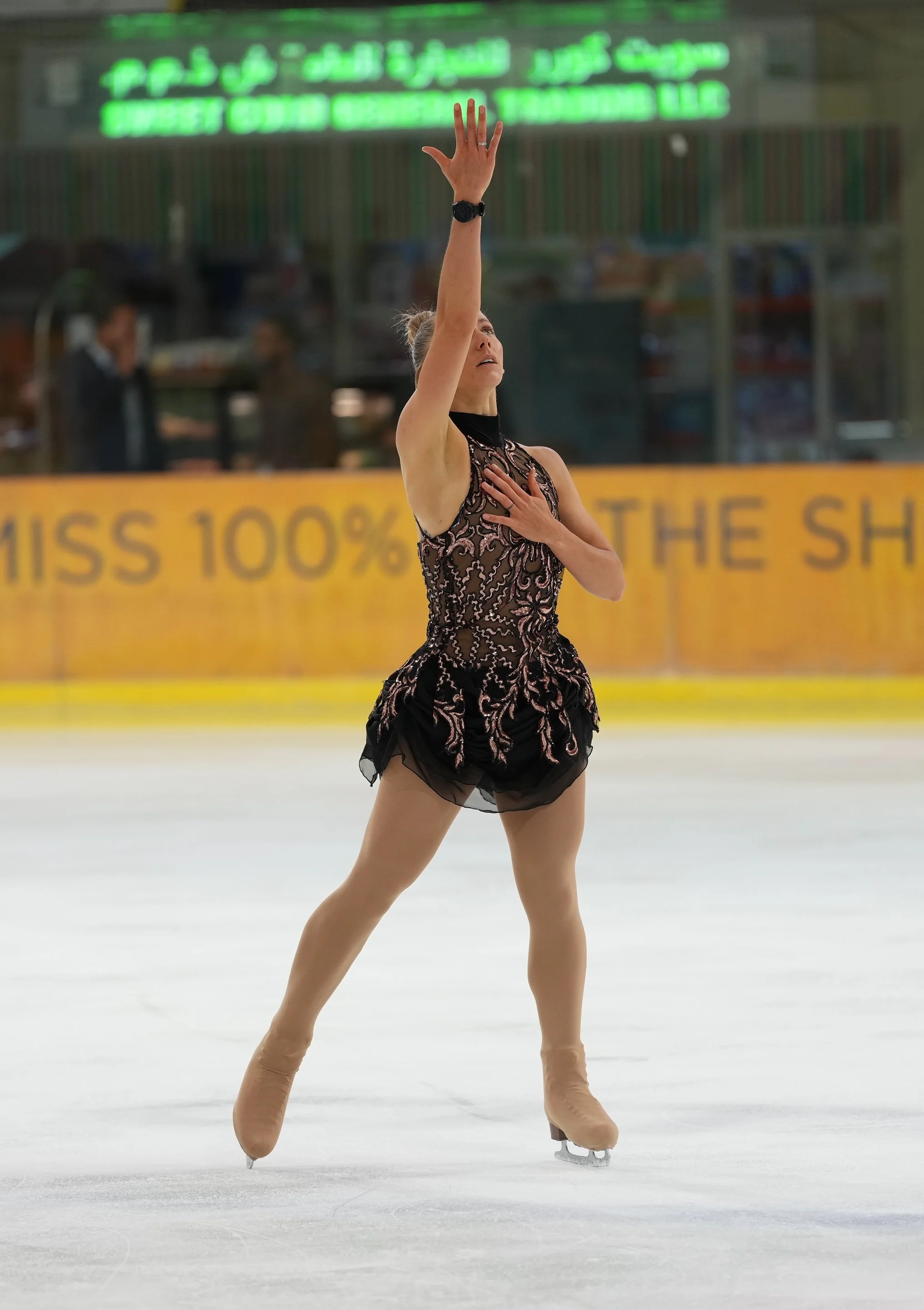 A female figure skater performing a move on the ice rink, wearing a black and pink dress, with one arm raised and the other hand on her chest.