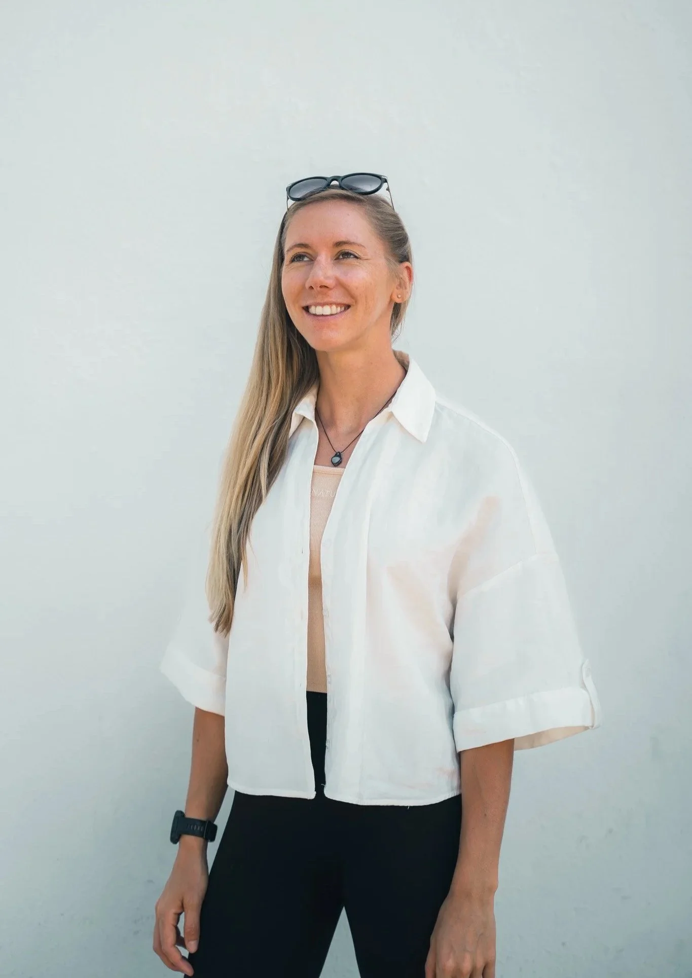 A woman with long blonde hair wearing a white short-sleeved shirt, black pants, sunglasses on her head, a watch on her left wrist, and a necklace, standing in front of a plain light-colored wall and smiling.