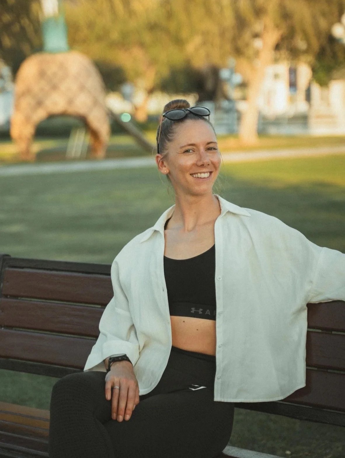 A woman sitting on a park bench smiling, wearing a black sports bra, white button-up shirt, black leggings, and sunglasses on her head, with a blurred outdoor park background with trees and a large abstract sculpture.