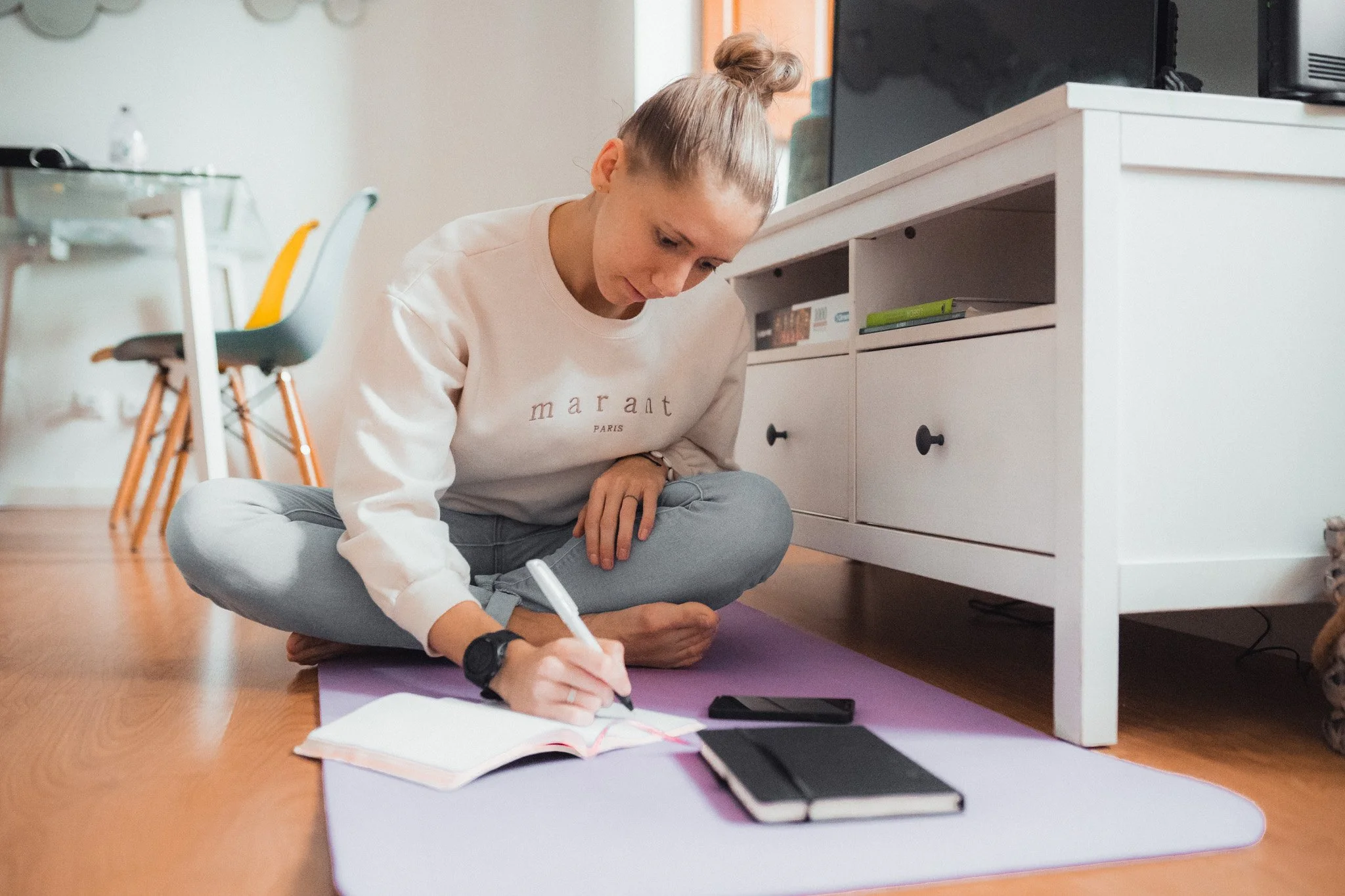 A woman practicing yoga indoors on a purple mat, sitting cross-legged and writing in a notebook, with a white cabinet and a television behind her.