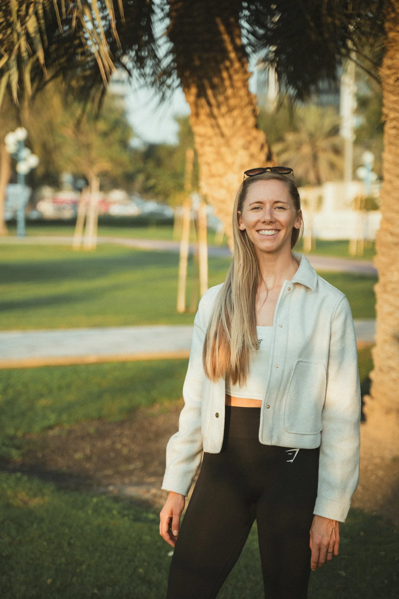 A young woman smiling outdoors during golden hour. She has long blonde hair, is wearing sunglasses on her head, a light-colored jacket, a white top, and black pants. She stands near palm trees in a park or similar setting.