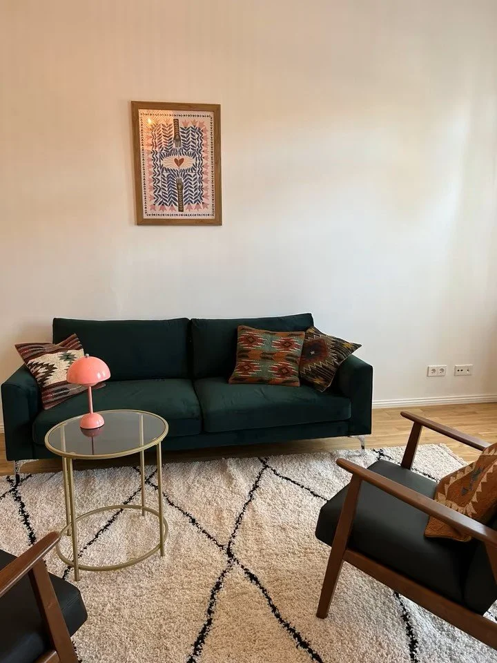 das Behandlungszimmer von Frau Dr. Sally Di Maio hat eine grüne Couch mit Kissen und einem kleinen Beistelltisch. Gegenüber ist ein Stuhl für Dr. Sally Di Maio der zum Sofa hin ausgerichtet ist