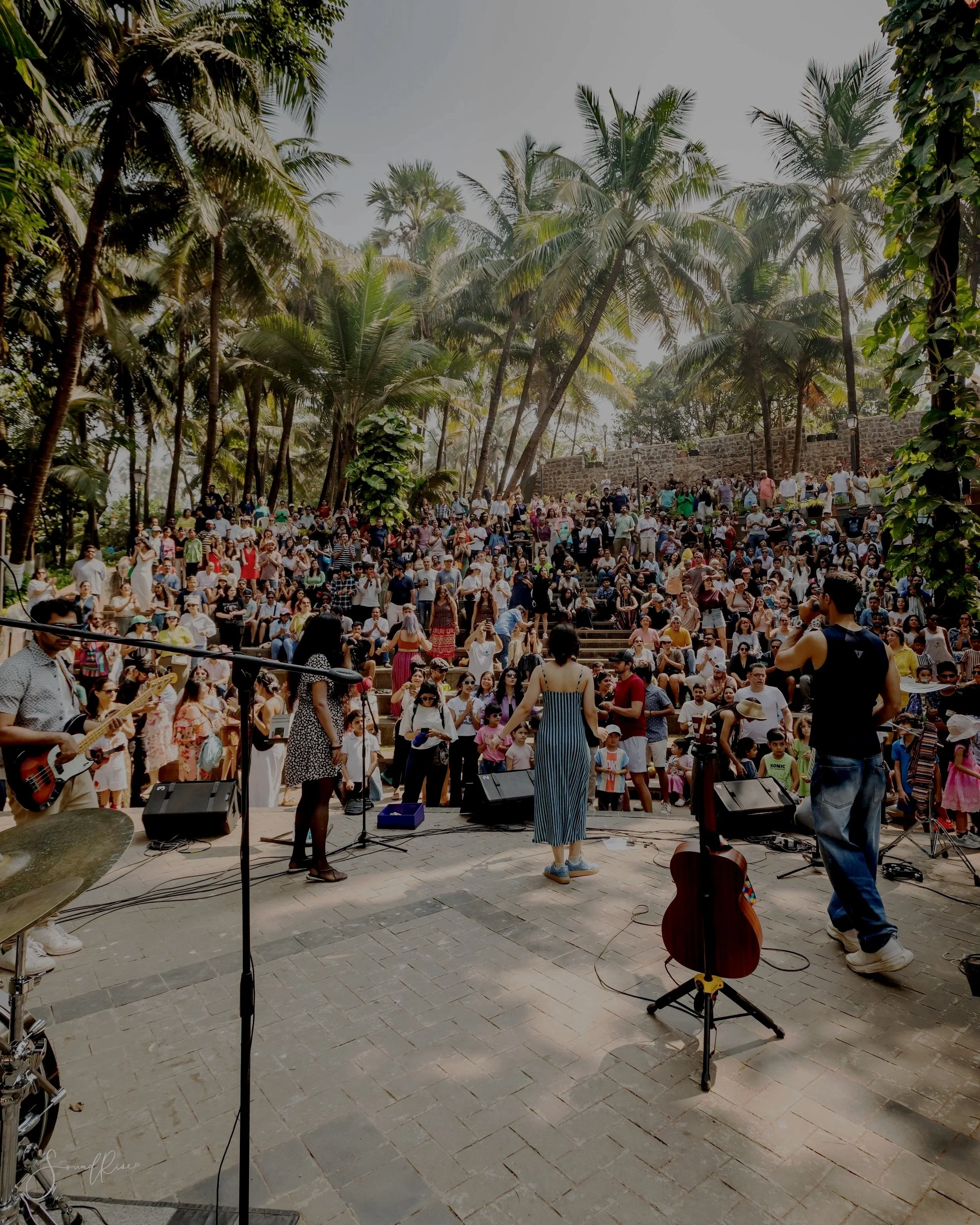 A large crowd gathered outdoors on stone steps for a concert or event in a lush, tropical setting with tall palm trees under a hazy sky. People are sitting, standing, and watching a performance on stage.