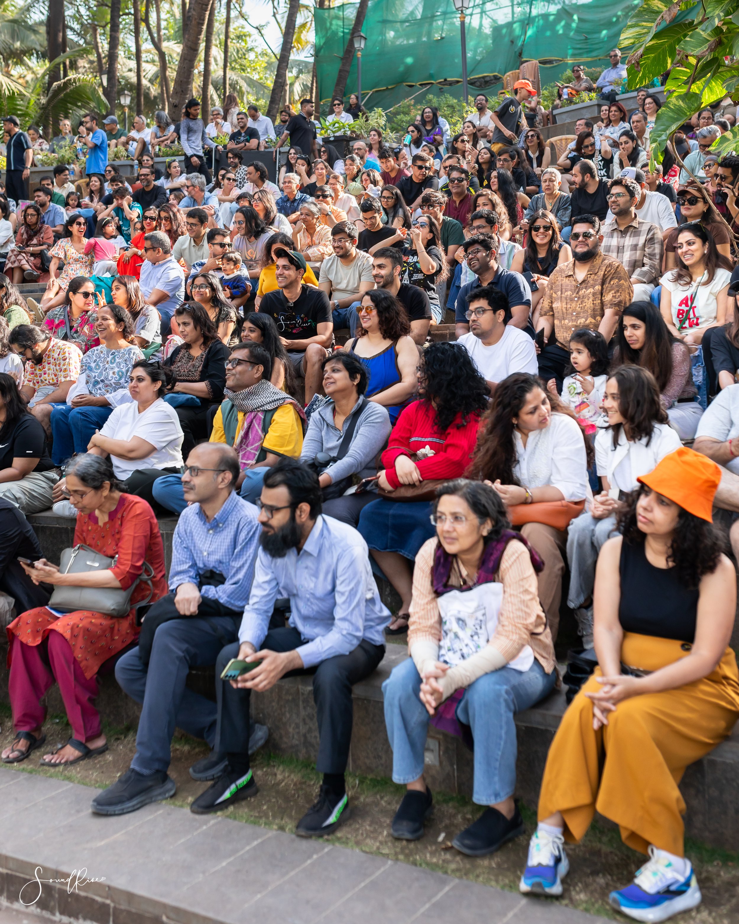 Crowd of diverse people sitting on outdoor tiered seating with trees and greenery in the background.