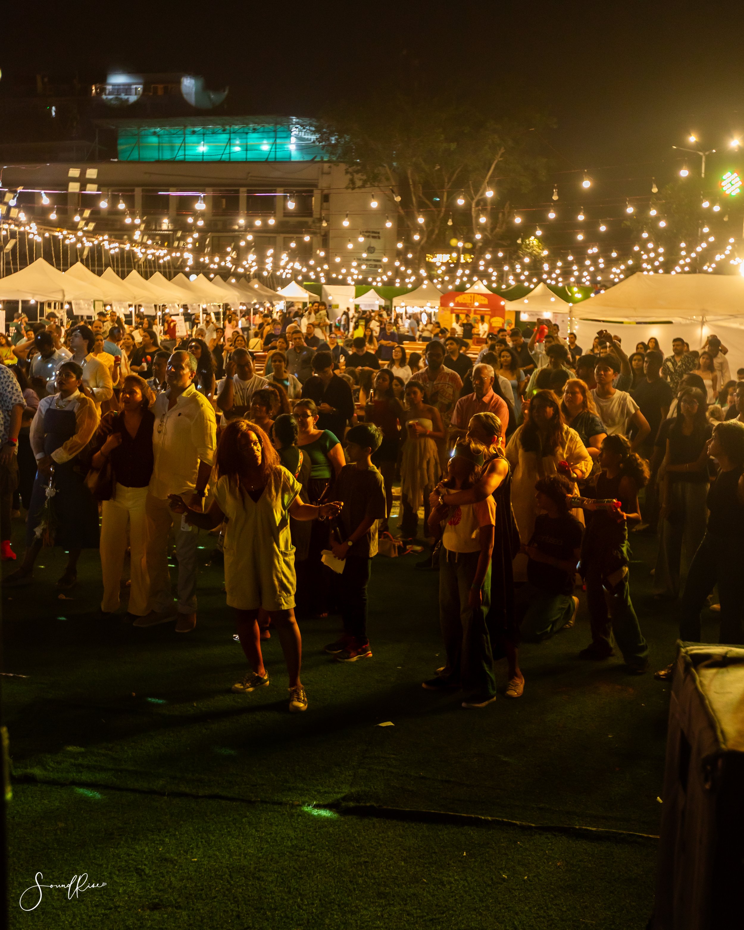 Night scene of a crowded outdoor event with string lights hanging overhead, numerous people standing and walking on a grassy area, and tents with vendor booths in the background.