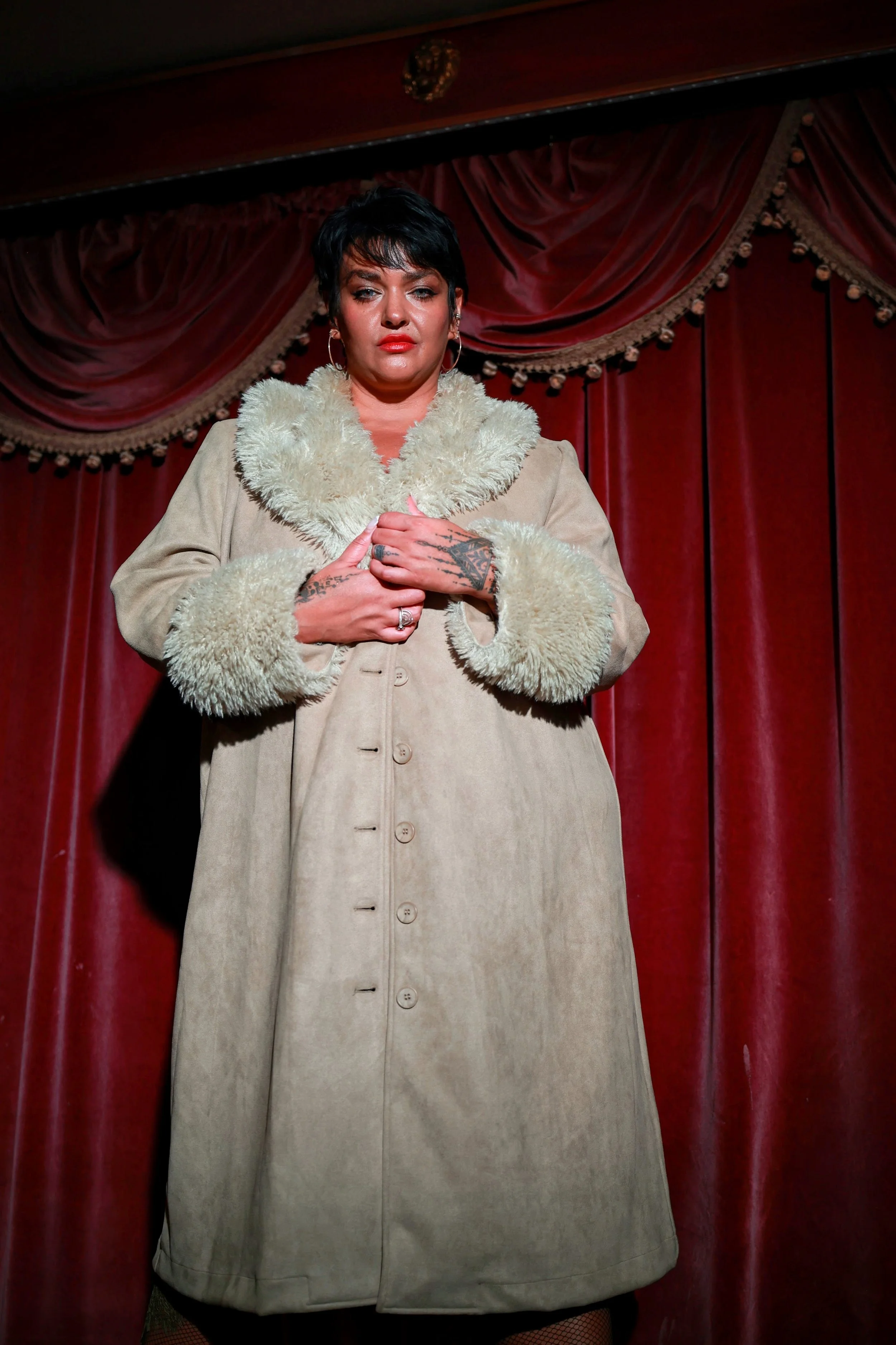 A person with short dark hair, wearing a beige coat with a fur collar, standing in front of a red curtain with gold trim, holding their hands to their chest. The person has visible tattoos on their hands.
