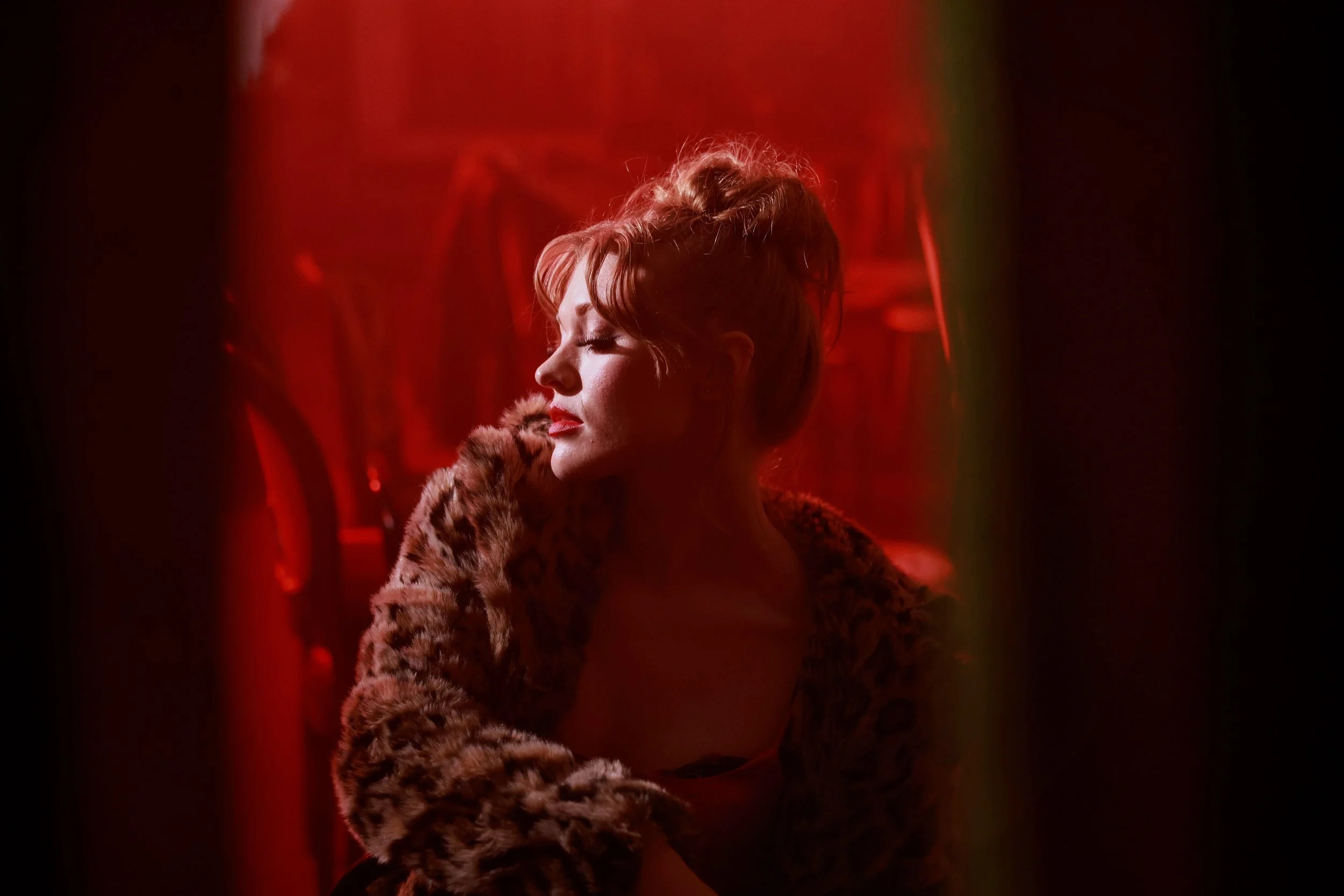 A woman with red hair in a bun, wearing a leopard print coat, in dim red lighting, with her eyes closed and a contemplative expression.