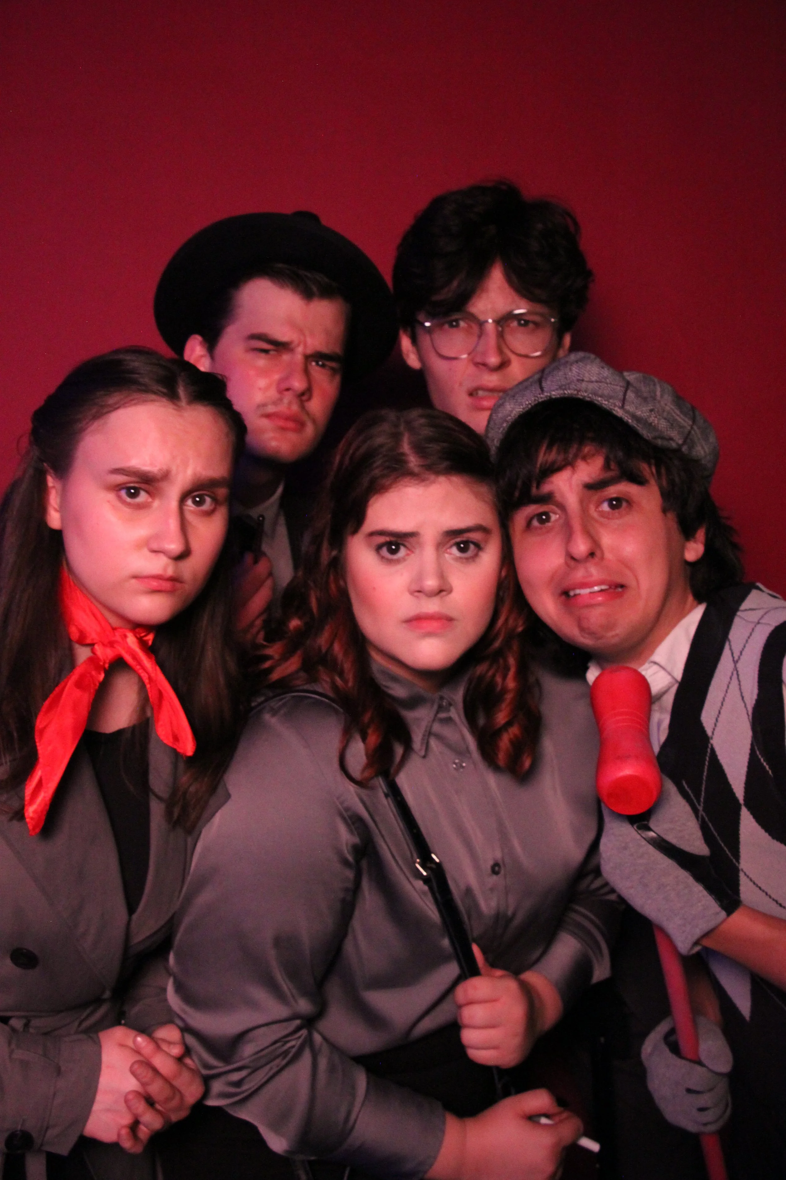 Group of young people with serious and confused expressions posing together against a red background, some wearing costumes and accessories.