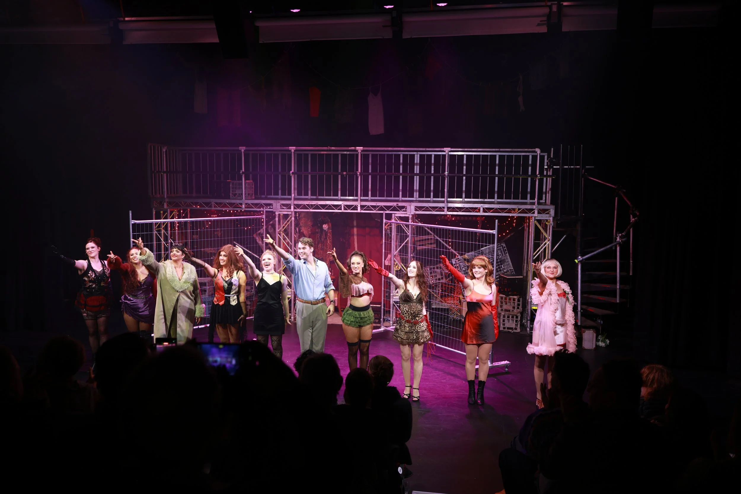 The image shows a stage with a cast of performers in costume, holding hands and taking a bow at the end of a theatrical performance. The background includes a theatrical set with scaffolding and various props.