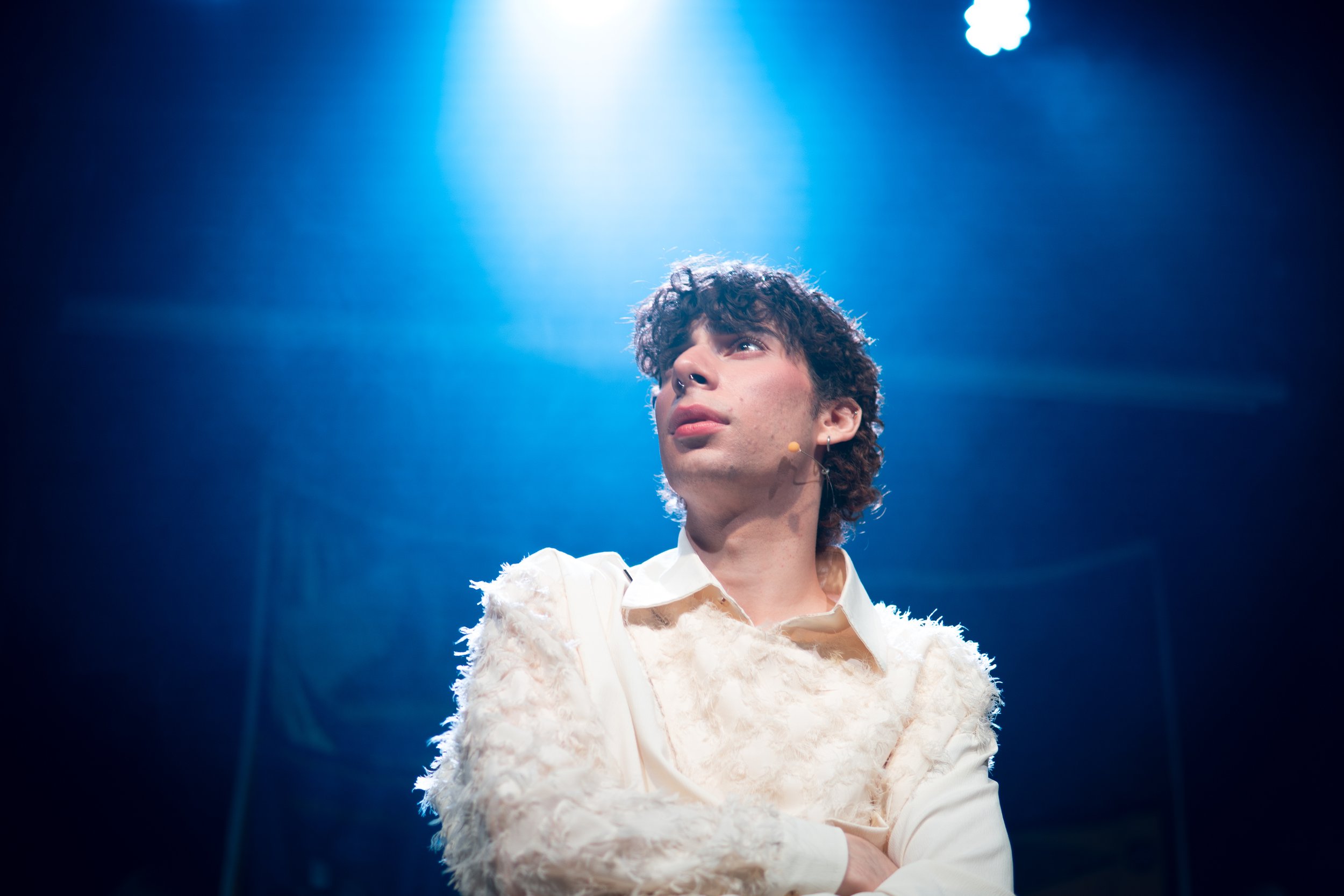 A young man with curly brown hair and piercings looks upward in a spotlight on stage, wearing a white ornate jacket.