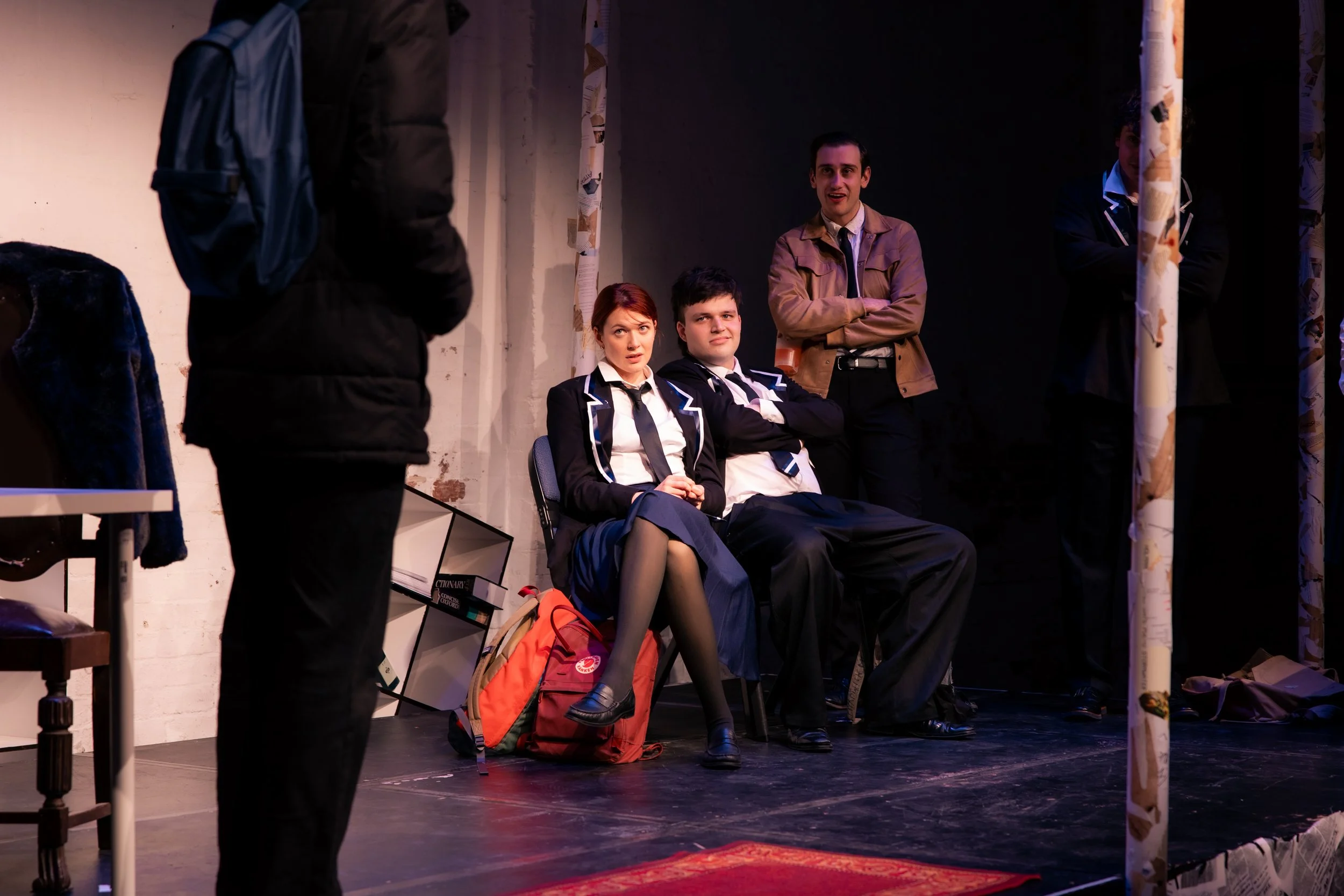 Four people, two women and two men, sitting and standing backstage at a theater or performance venue, with various belongings such as backpacks and shoes around them. They are engaged in conversation or waiting, with one person looking at the others. The setting appears informal and behind the scenes.