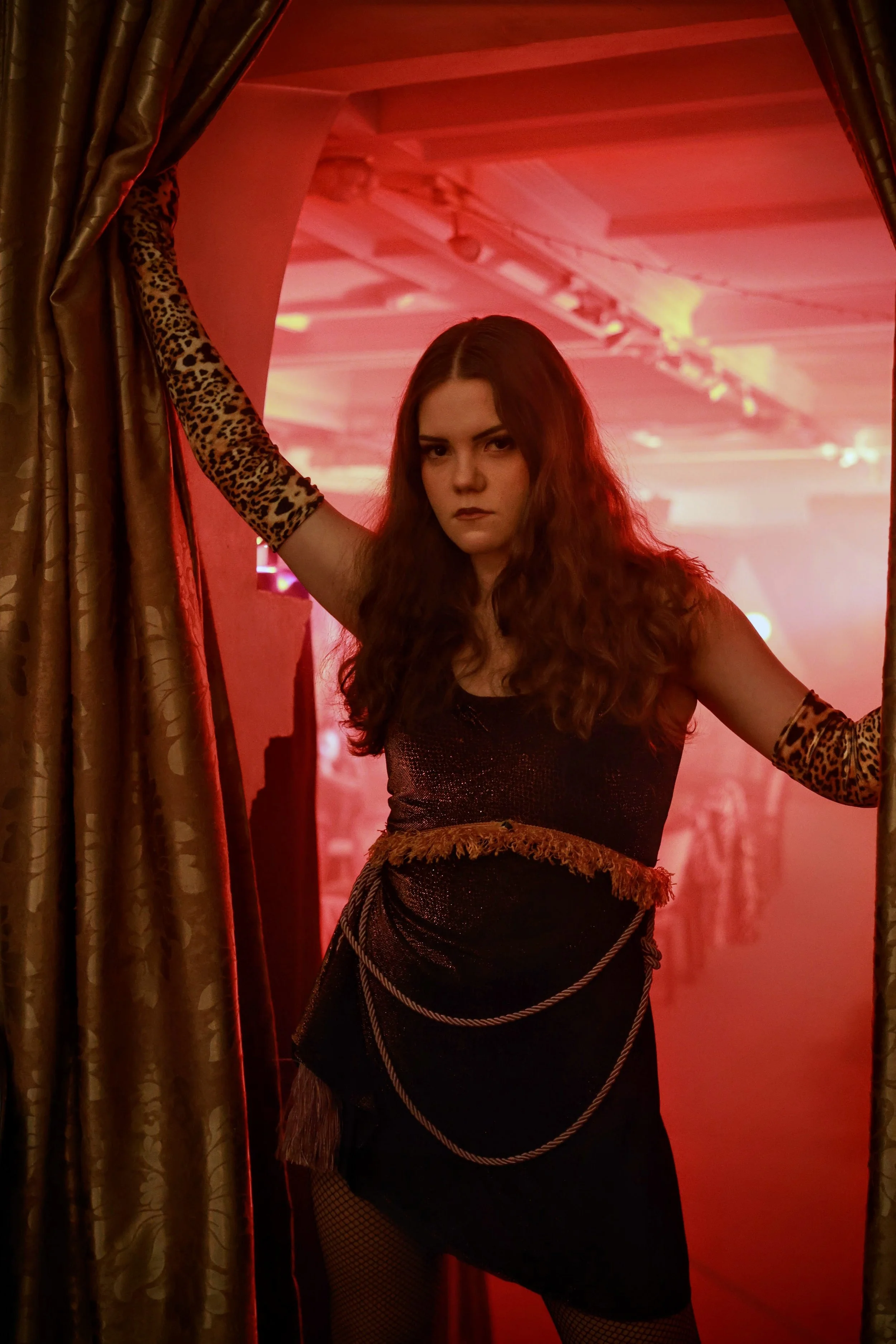 A woman with long, wavy hair stands in front of a red-lit background. She has a serious expression, wears a black dress, leopard print gloves, fishnet stockings, and a chain belt. She is holding a curtain with her left hand, and her right arm rests on her hip.