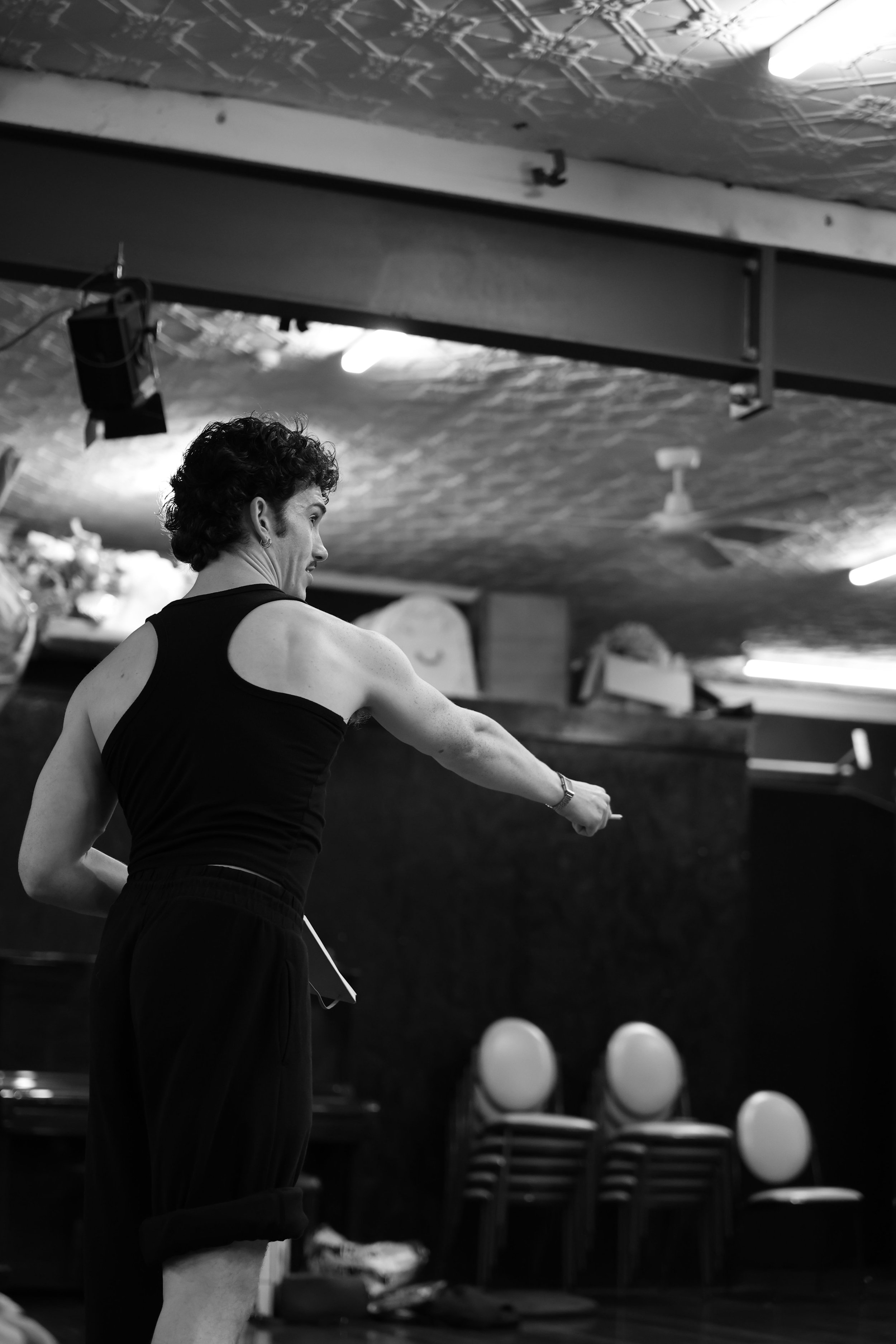 A black-and-white photo of a choreographer with short, curly hair wearing a sleeveless top and large earrings, standing indoors in a room with stacked chairs in the background, holding a script and directing unseen dancers