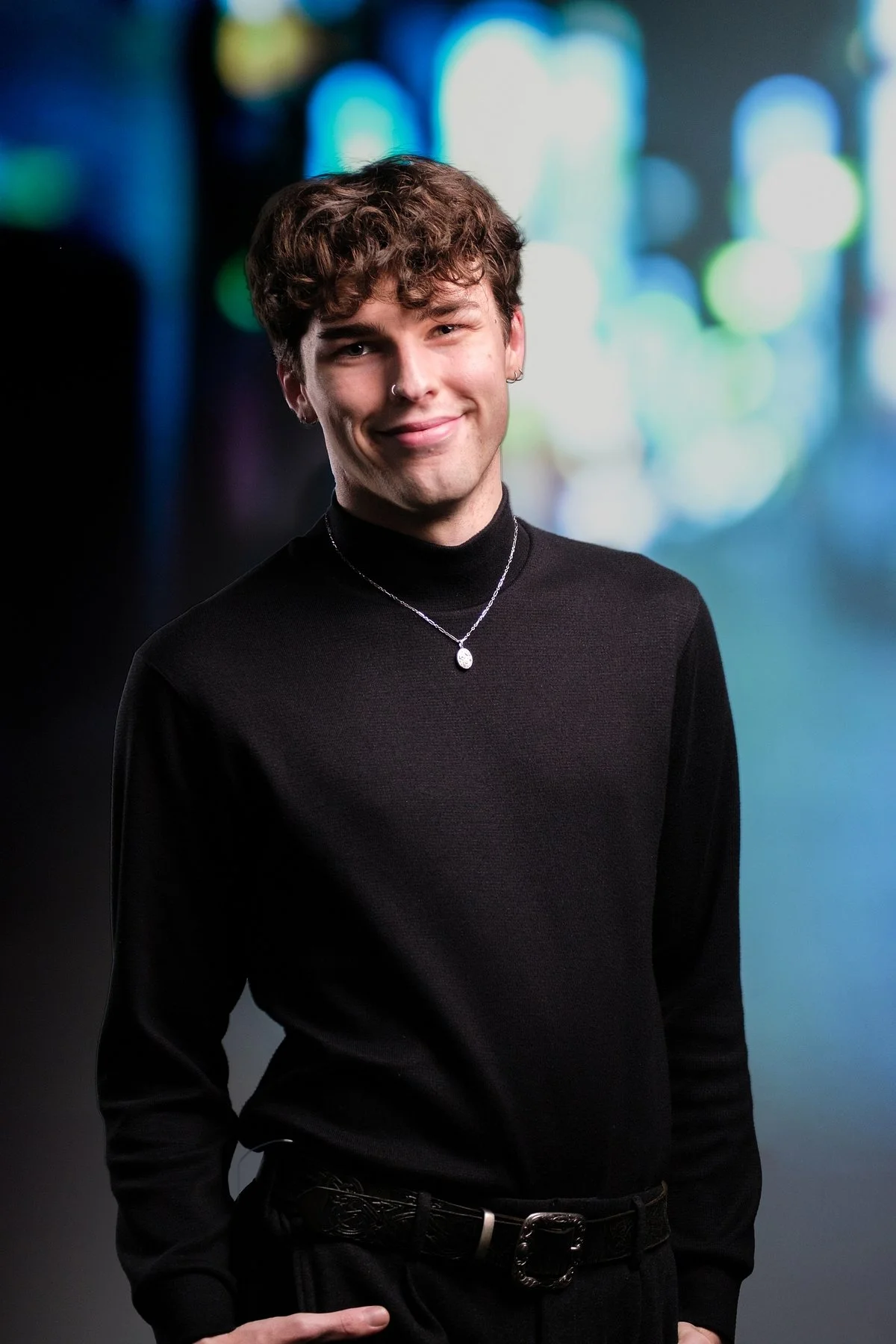 A young person with curly brown hair, wearing a black turtleneck sweater and a necklace, smiling at the camera against a blurred, colorful bokeh background.