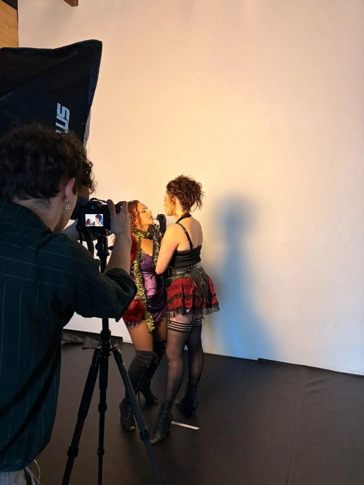 Two women in colorful, edgy outfits pose for a photo in front of a plain white wall while a photographer captures the moment.