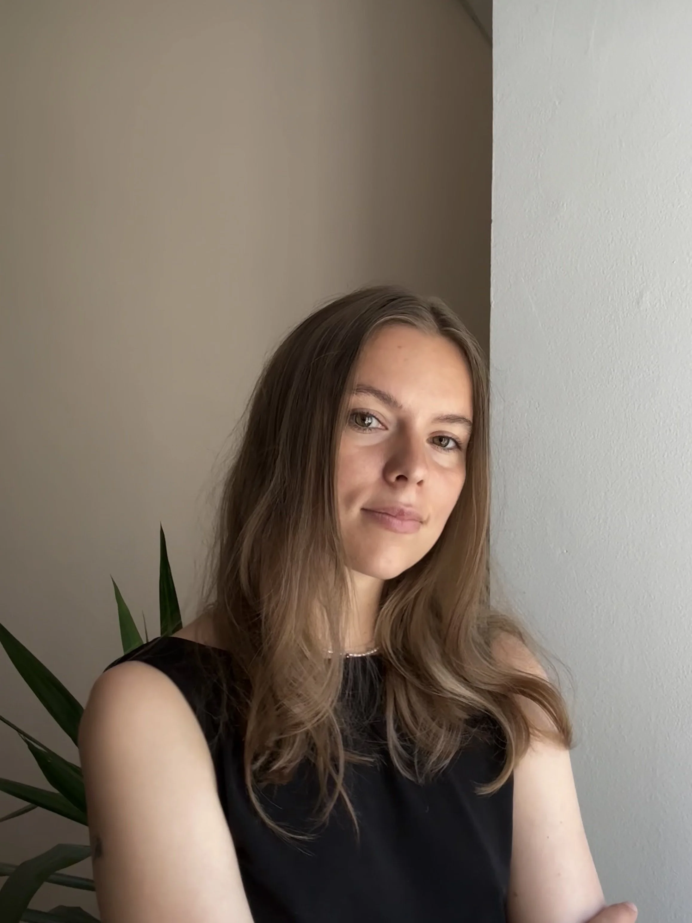 A young woman with long, wavy brown hair, wearing a black sleeveless top, smiling slightly while standing indoors near a white wall and some green houseplants.