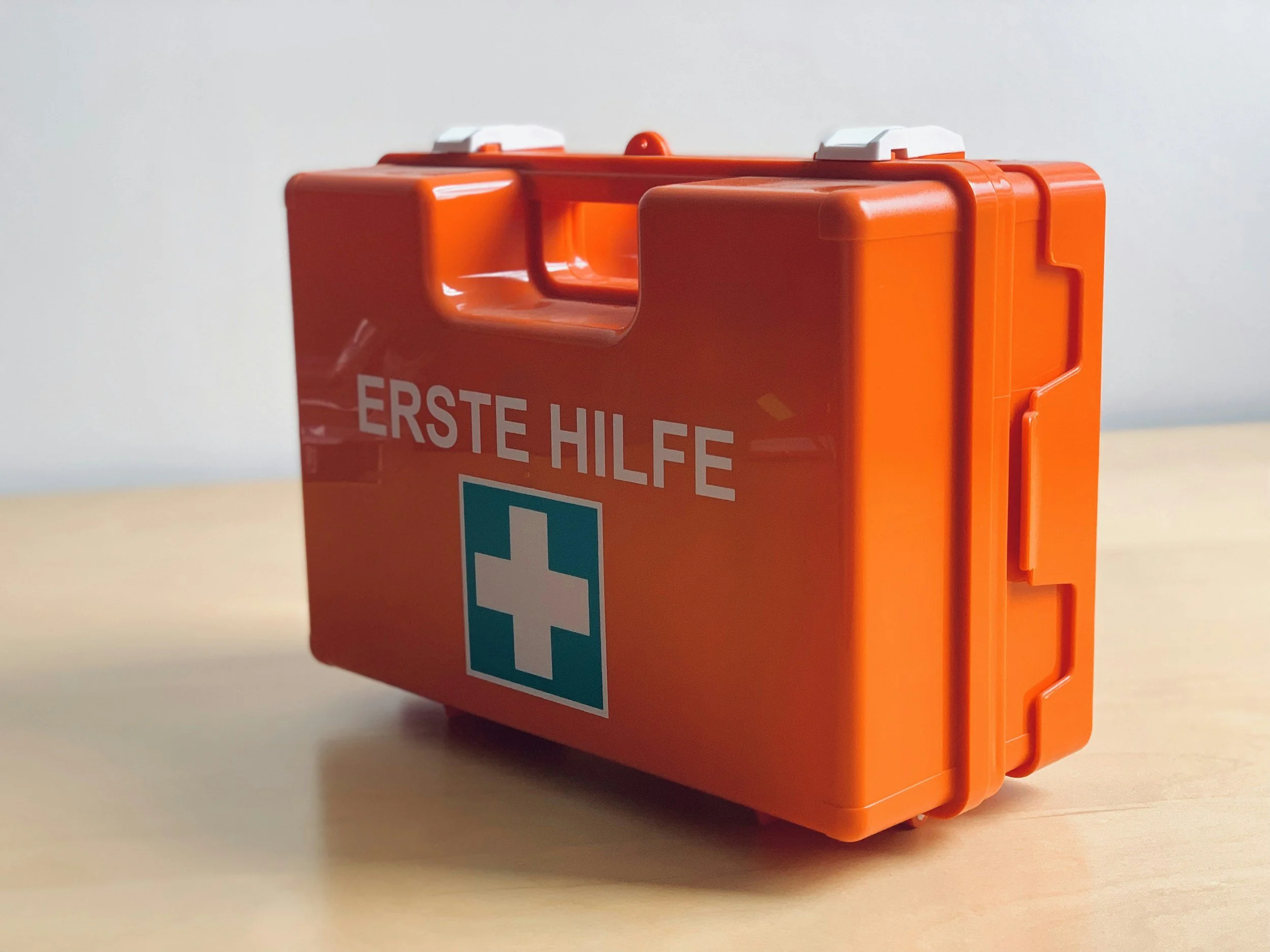 Ein rotes Erste-Hilfe-Koffer mit weißem Kreuz und blauer Markierung steht auf einem Tisch gegen eine weiße Wand.