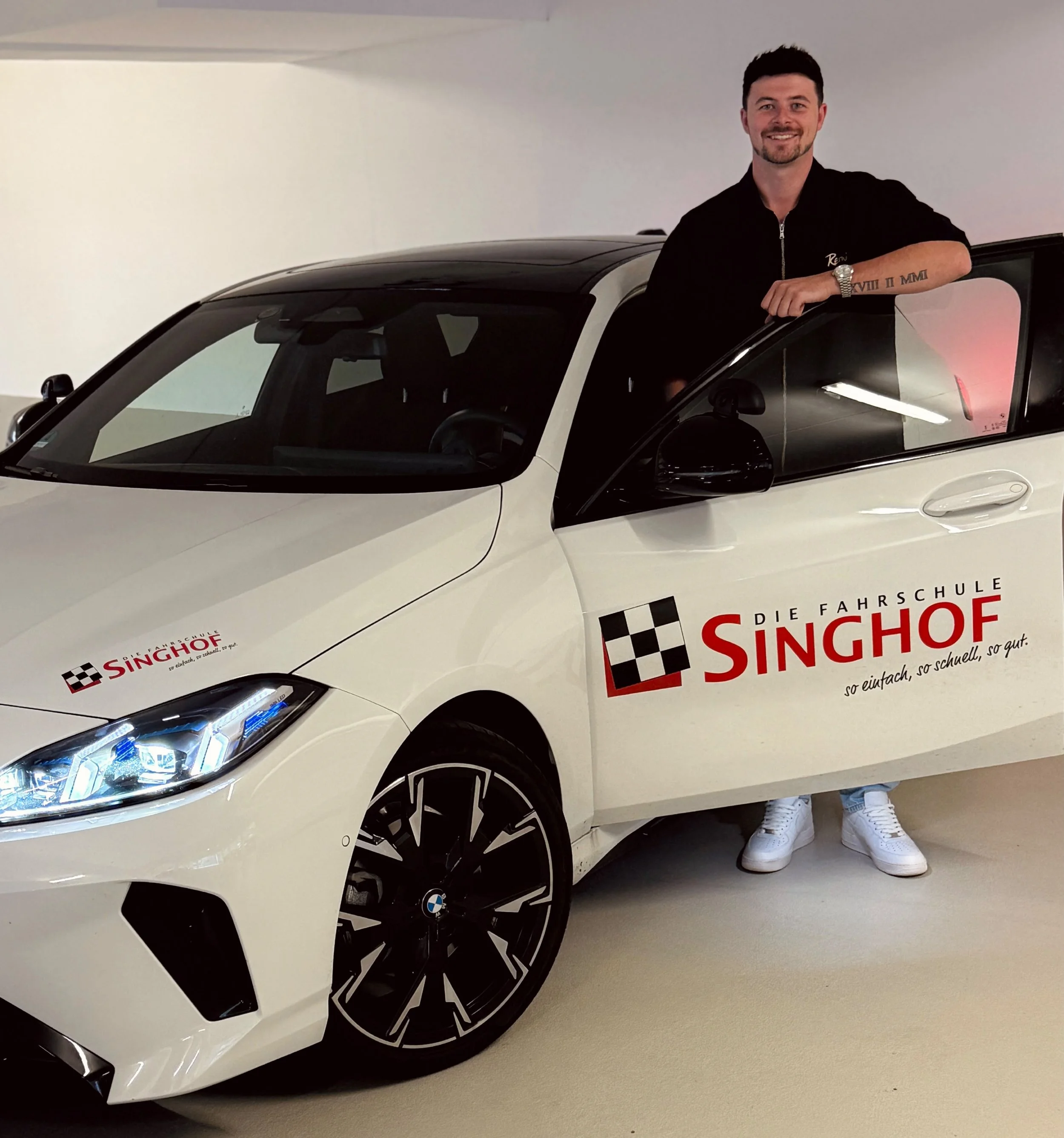 Junge Mann steht vor weißem Auto mit Singhof-Logo auf einem Parkplatz, im Hintergrund Bäume und Fahrräder.