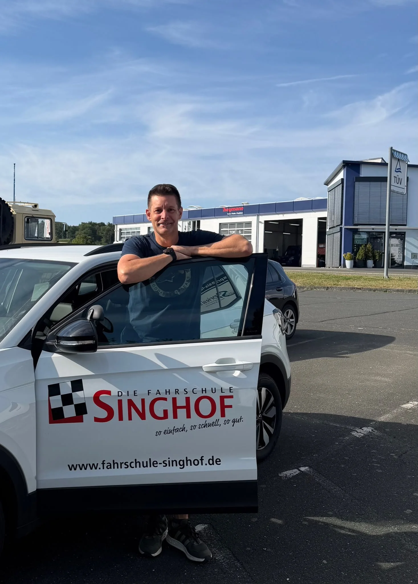 Ein Mann sitzt im Auto, trägt ein weißes T-Shirt und lächelt in die Kamera.