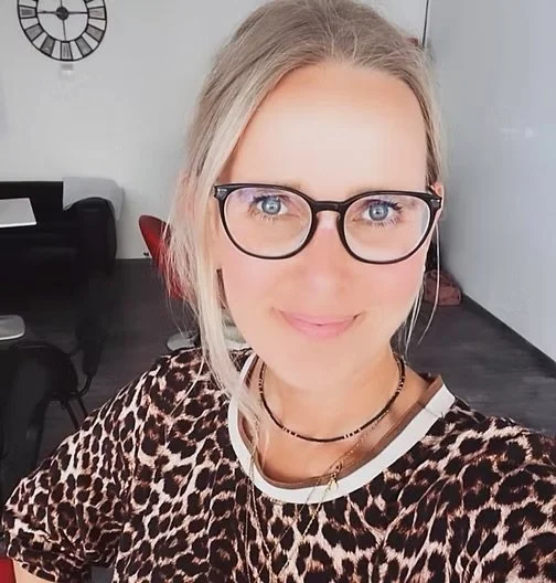 Junge Frau mit Brille und blonden Haaren, trägt ein Leopardenmuster-Shirt, im Hintergrund ein Zimmer mit Uhr an der Wand.