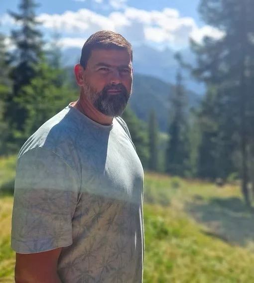 Ein Mann mit Bart und kurzen Haaren steht in einer Naturkulisse mit Bäumen und Bergen im Hintergrund, bewölktem Himmel, bei Sonnenlicht.