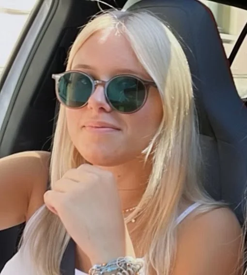 Junge Frau mit blonder Haar, Sonnenbrille und weißen Oberteil sitzt im Auto.