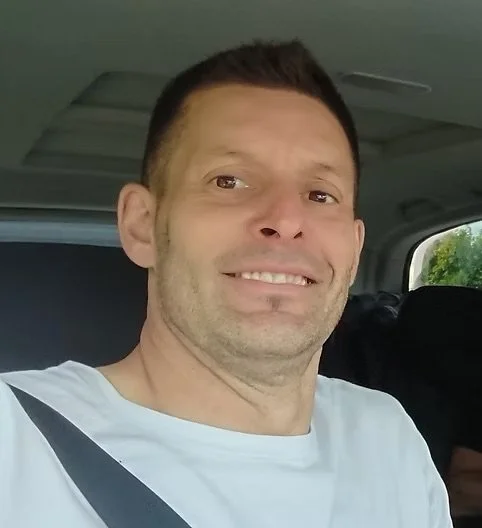 Ein Mann sitzt im Auto, trägt ein weißes T-Shirt und lächelt in die Kamera.