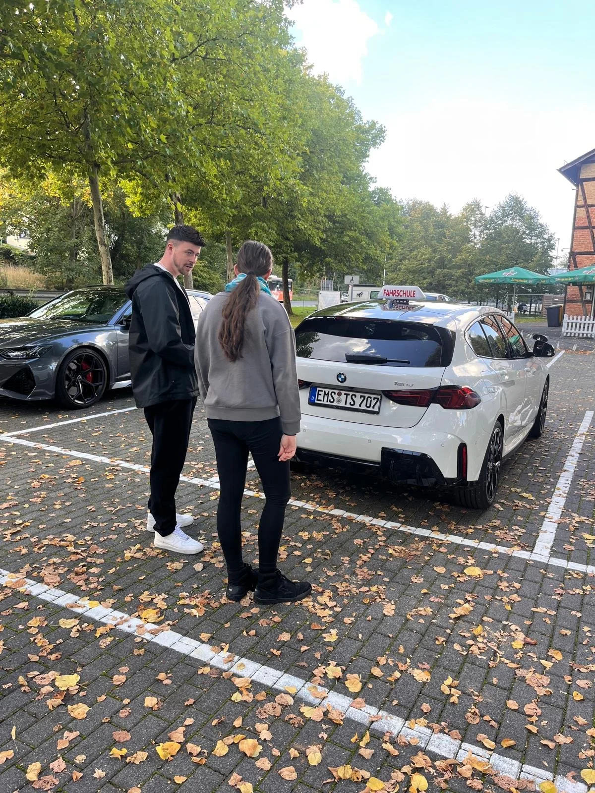 Zwei Menschen sprechen in einem Parkhaus. Es gibt ein weißes Auto mit einem Fahrschullogoschild auf dem Dach. Der Mann trägt schwarze Kleidung und weiße Schuhe, das Mädchen trägt schwarze Schuhe, schwarze Leggings und eine graue Jacke.