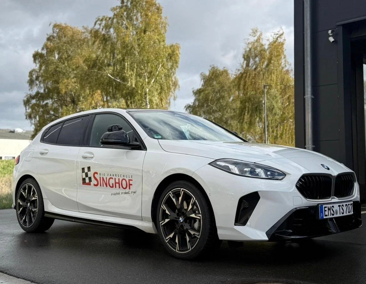 Ein weißes Auto mit dem Logo der Fahrschule Singhof, das vor einer modernen Gebäudefassade steht, umgeben von Bäumen mit herbstlichen Blättern.
