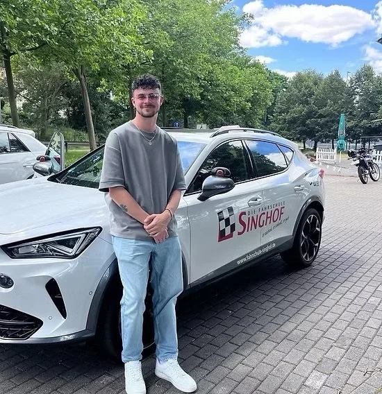 Junge Mann steht vor weißem Auto mit Singhof-Logo auf einem Parkplatz, im Hintergrund Bäume und Fahrräder.