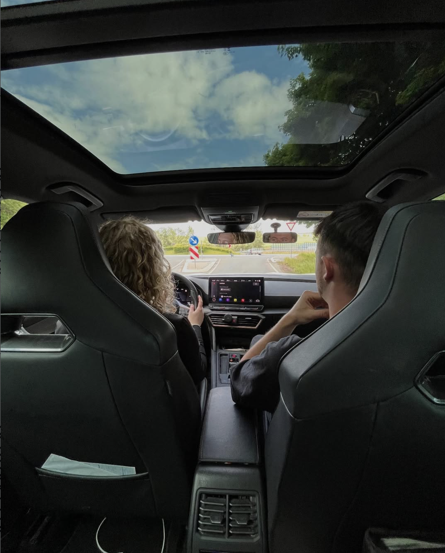 Innenansicht eines Autos, zwei Personen sitzen auf den Vordersitzen, die Beifahrerin, eine Frau mit lockigem Haar, fährt, während der Fahrer, ein Mann, auf das Lenkrad schaut. Das Display im Auto zeigt eine Navigationskarte. Das Dach ist eine große Sonnenluke, die den Himmel mit Wolken zeigt. Außen sind Bäume und eine Straße sichtbar.