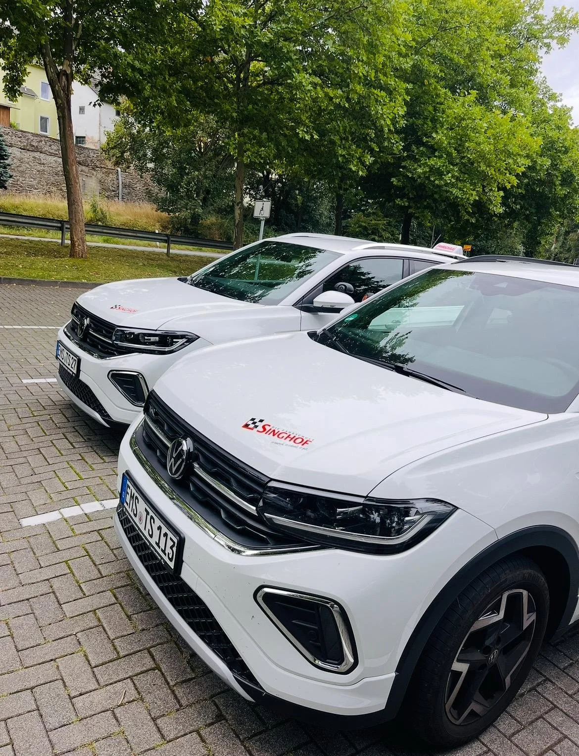Zwei weiße Autos mit Singhof- und VW-Logos parken auf einem Parkplatz, umgeben von Bäumen und einem Zaun, in einer grünen, urbanen Umgebung.