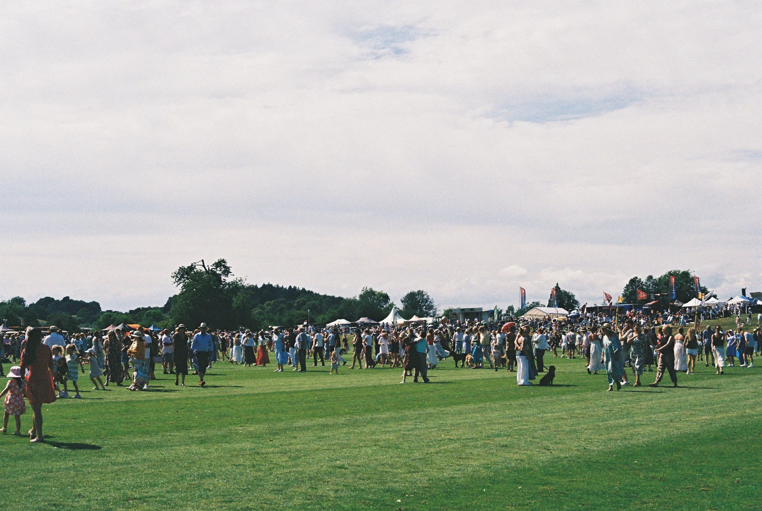 A large outdoor event with many people gathered on a grassy field, tents and small stages in the background, and an overcast sky.