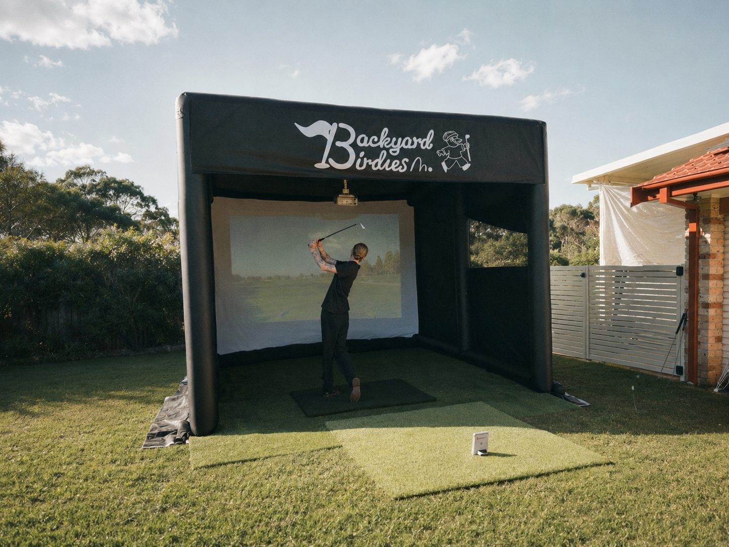Person practicing golf swing inside an outdoor simulator with a screen, under a black canopy labeled "Backyard Birdies".