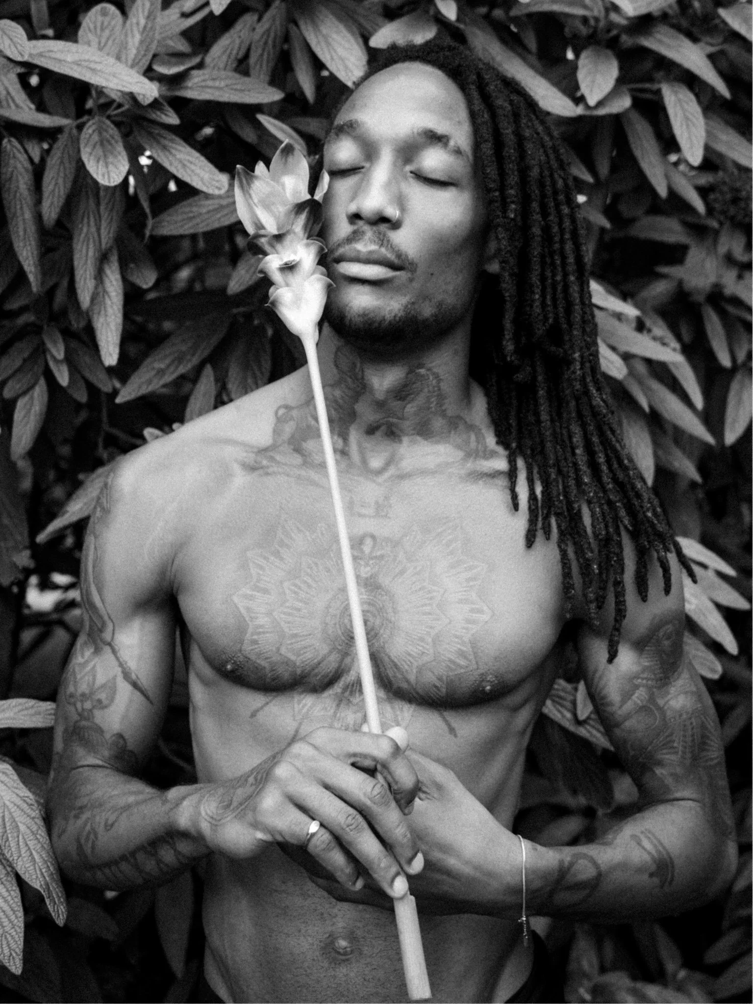 Black and white portrait of a tattooed man holding a flower among dense foliage with eyes closed.