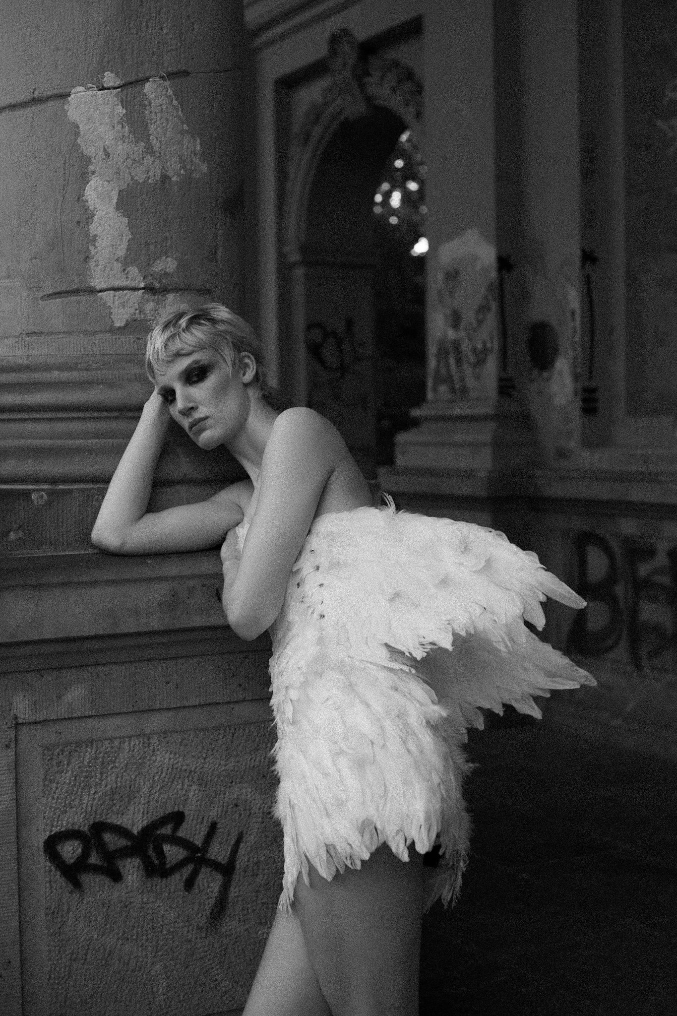 A woman with short hair and makeup leaning against a wall in an indoor setting, wearing a dress with large feathered wings attached.