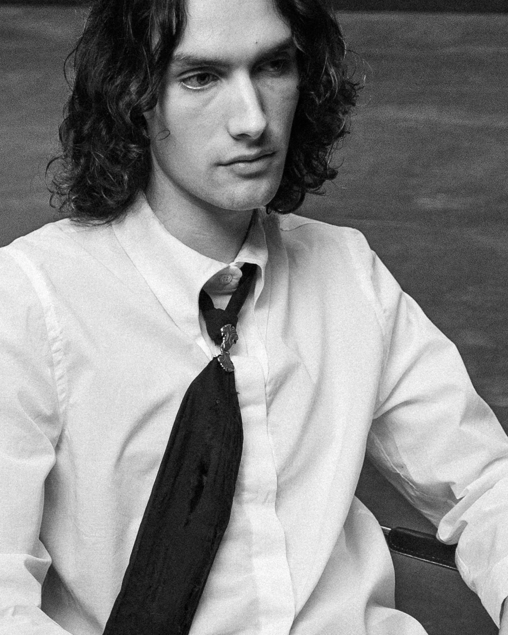 Black and white photograph of a person with shoulder-length wavy hair, wearing a collared shirt, and a dark tie, looking downward with a neutral expression.
