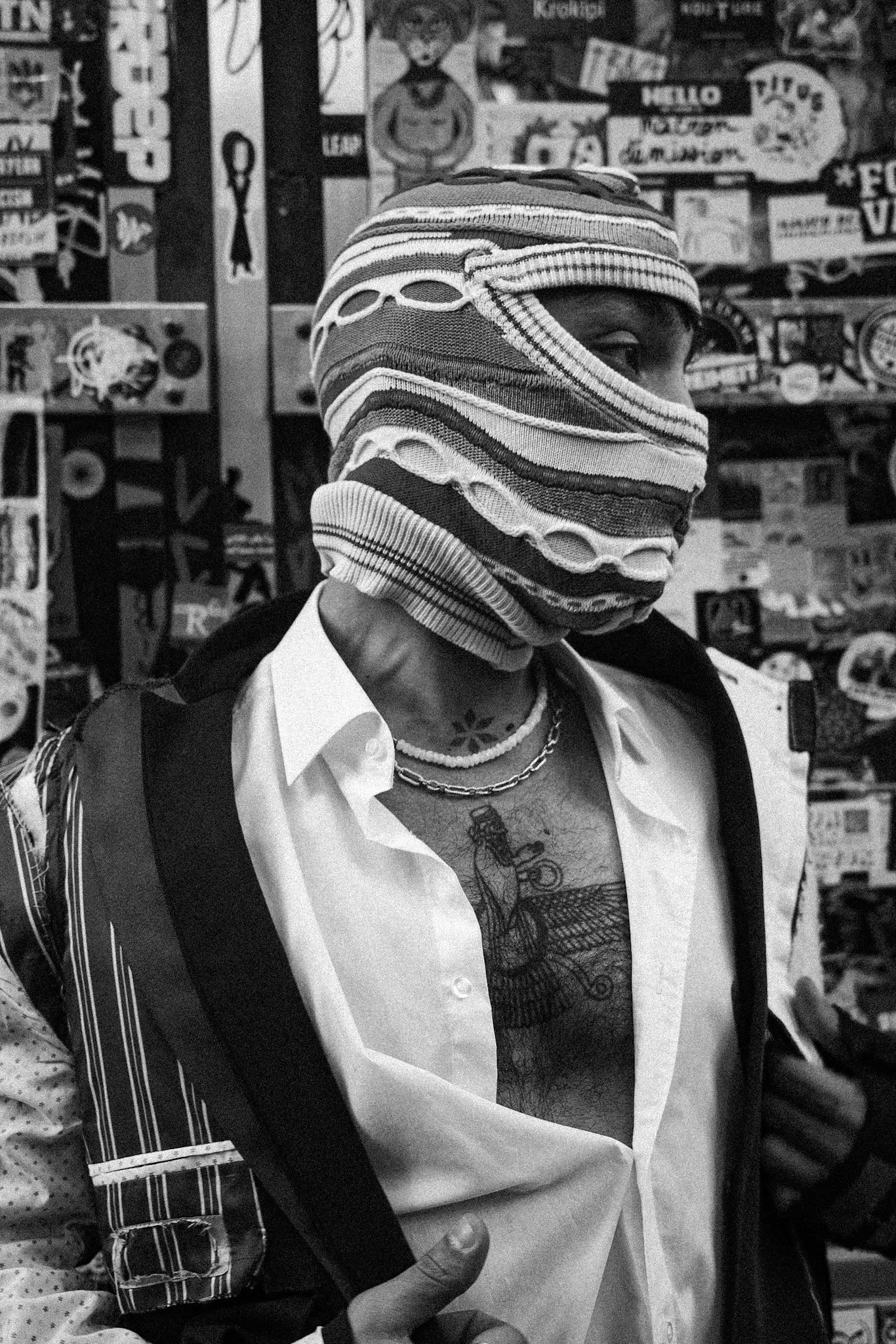 Street portrait of a person wearing a knitted face covering in front of a sticker-covered wall in Berlin. Black and white photograph.
